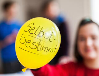 Jugenclub BlueBox Selbstbestimmung Zu sehen ist ein gelber Luftballon, auf dem "selbstbestimmt" steht. Im Hintergrund sieht man verschwommen eine junge Person, die den Luftballon hält.
