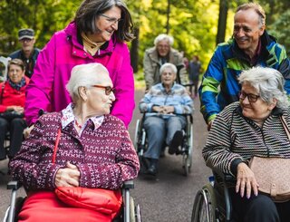 Mobilitätshilfedienst Mitte Gemeinsamer Ausflug Zu sehen sind von vorner zwei ältere Frauen in Rollstühlen. Sie schauen sich lächelnd an. Gefahren werden sie ebenfalls von zwei Personen, die lächeln und ihre Blicken ihnen zugwandt haben. Sie befinden sich auf einem Weg in einem sehr grünem Gelände. Im Hintergrund sind weitere Personen in Rollstühlen zu sehen.