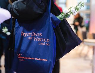 Humanistischer Verband Berlin-Brandenburg: Humanistische Hochschule Berlin Auf einem dunkelblauen Beutel, den eine Person über ihrer linken Schulter trägt, steht: „Irgendwas mit Werten uns so.“ Das Logo der Humanistischen Hochschule Berlin ist unten auf dem Beutel in Orange zu sehen.