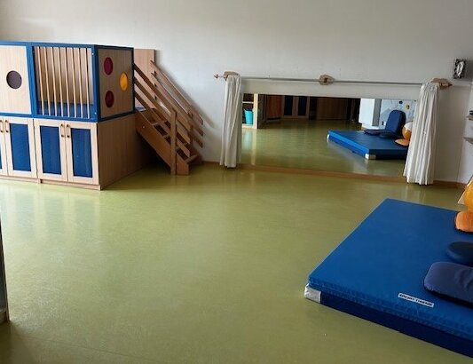 Auf einem Teppich mit Kinderpiratenmotiv steht ein Korb mit Kissen, dahinter ist die hohe Fensterfront zu sehen. Rechts steht ein Kleiderschrank. Links von ihm befindet sich eine Matratze mit zwei Kissen. Daneben steht ein Sessel mit einem Kissen.