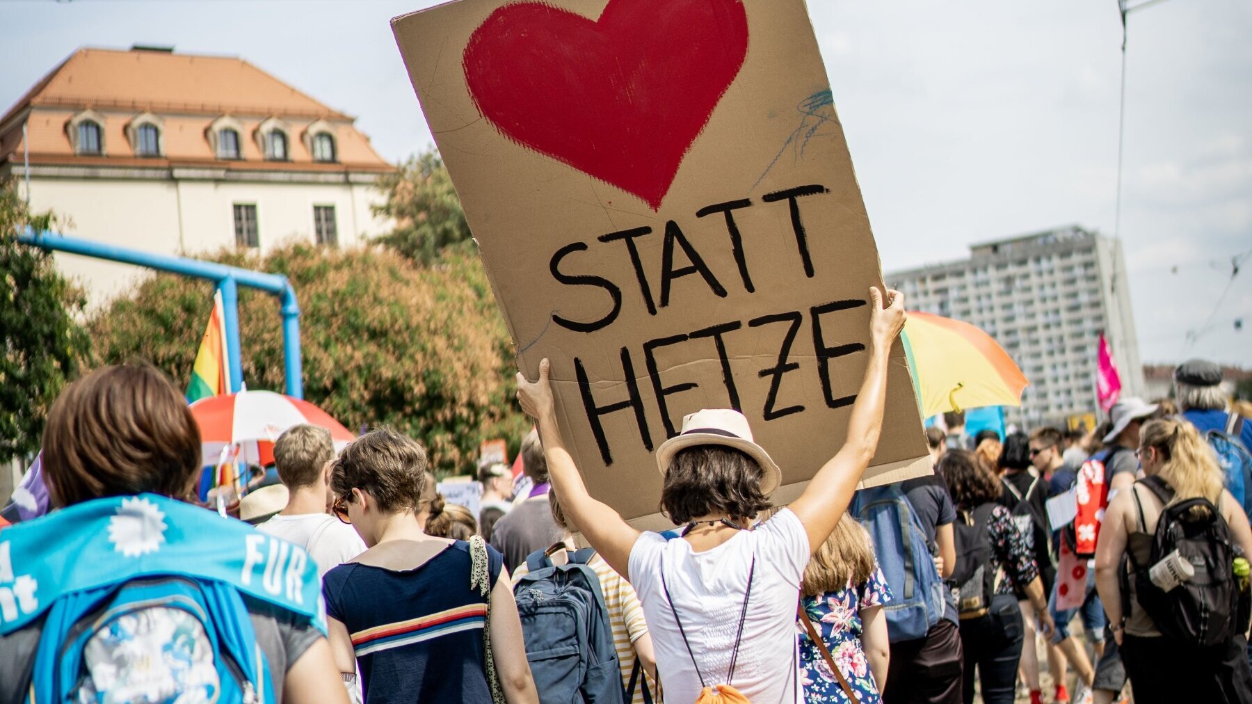 Demonstrierende, die von hinten zu sehen sind, laufen auf der Straße. Eine Person hält ein Pappschild hoch, auf dem ein rotes Herz zu sehen ist. Darunter steht in schwarzen Großbuchstaben: „statt Hetze“.