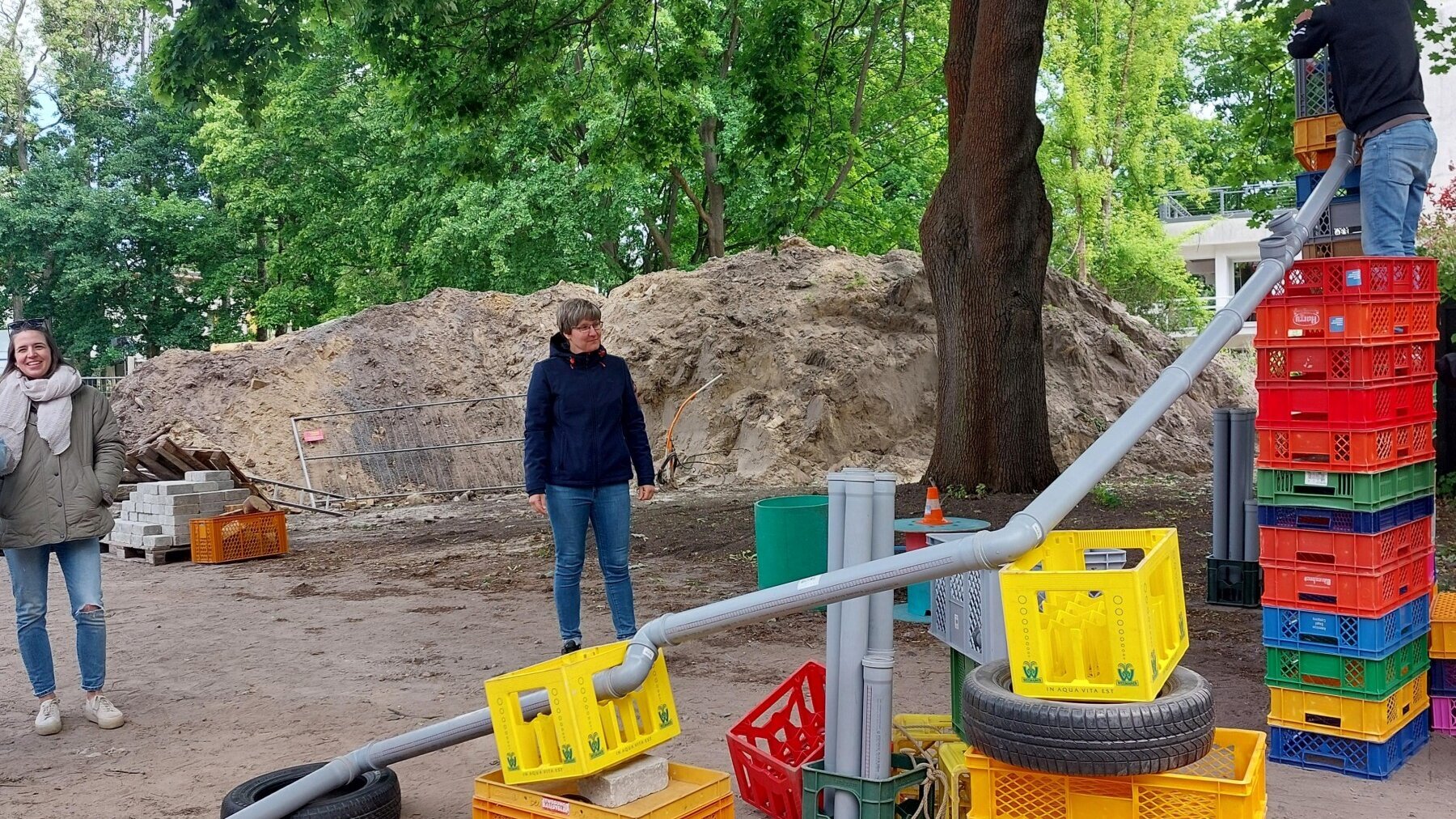 Draußen sind unterschiedlich hohe Kistentürme gestapelt, auf denen ein Plastikrohr liegt. Rechts stapelt ein Mann Kisten, zwei Frauen stehen hinter der Konstruktion.
