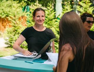 Humanistische Fachschule für Sozialpädagogik in Berlin: Angebote Eine Frau steht draußen an einem Tresen, ihre rechte Hand liegt auf einem geöffneten Ordner, mit der rechten hält sie den Ordnerdeckel. Rechts vor dem Tresen steht eine Frau und guckt sich ein DIN-A4-Blatt an.