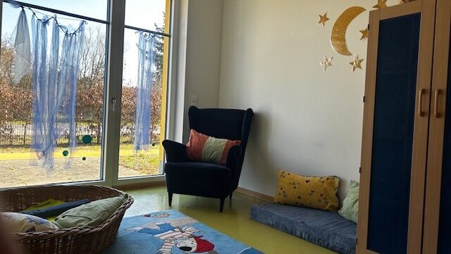 Auf einem Teppich mit Kinderpiratenmotiv steht ein Korb mit Kissen, dahinter ist die hohe Fensterfront zu sehen. Rechts steht ein Kleiderschrank. Links von ihm befindet sich eine Matratze mit zwei Kissen. Daneben steht ein Sessel mit einem Kissen.