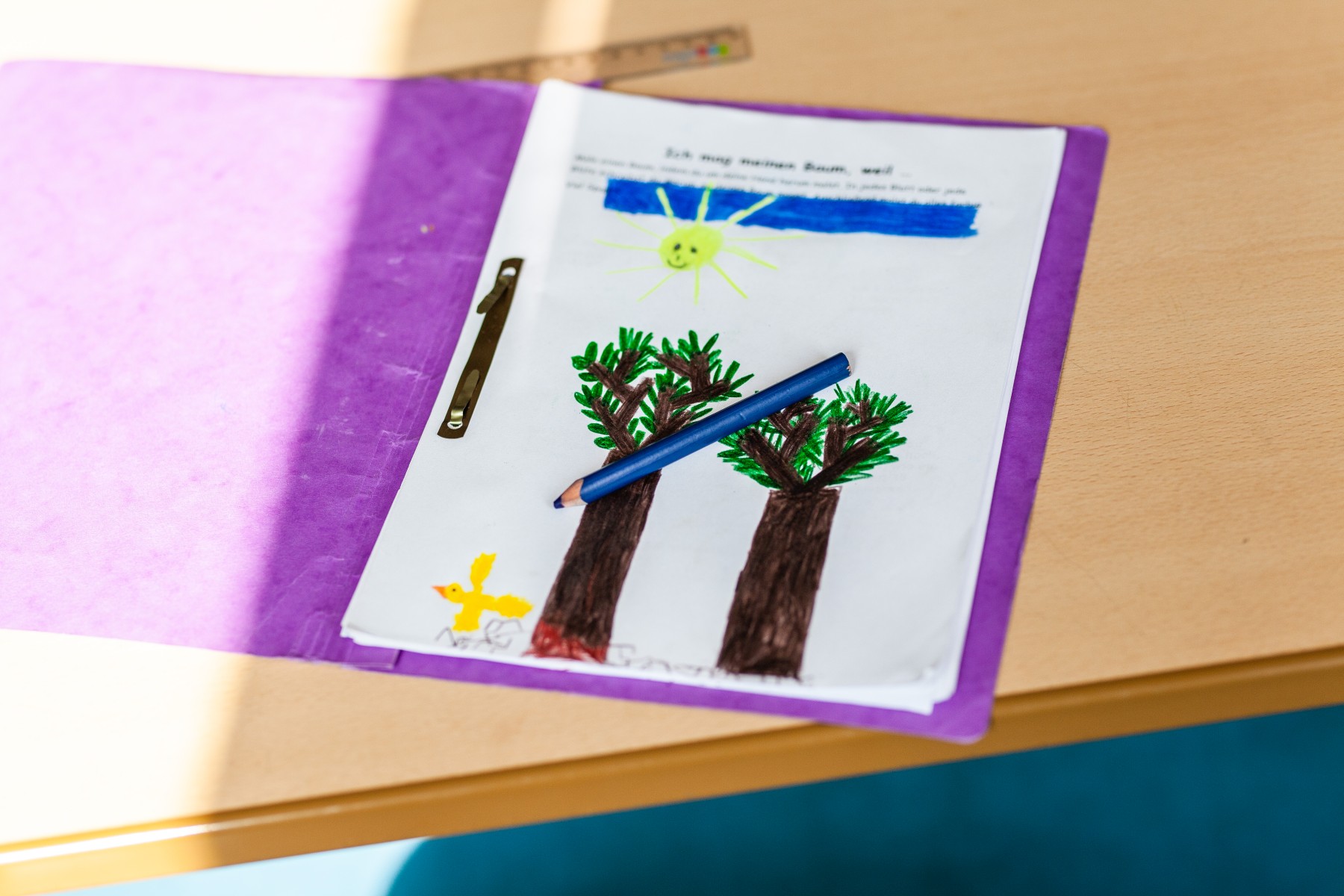 Auf einer Tischplatte liegt ein geöffneter lila Papphefter. Darin ist das obere Arbeitsblatt zu sehen. Darauf wurden ein Himmel, eine Sonne, zwei Bäume und links unten neben dem Baum links ein Vogel mit Filzstiften gemalt. Auf dem Blatt liegt quer ein dunkelblauer Buntstift.