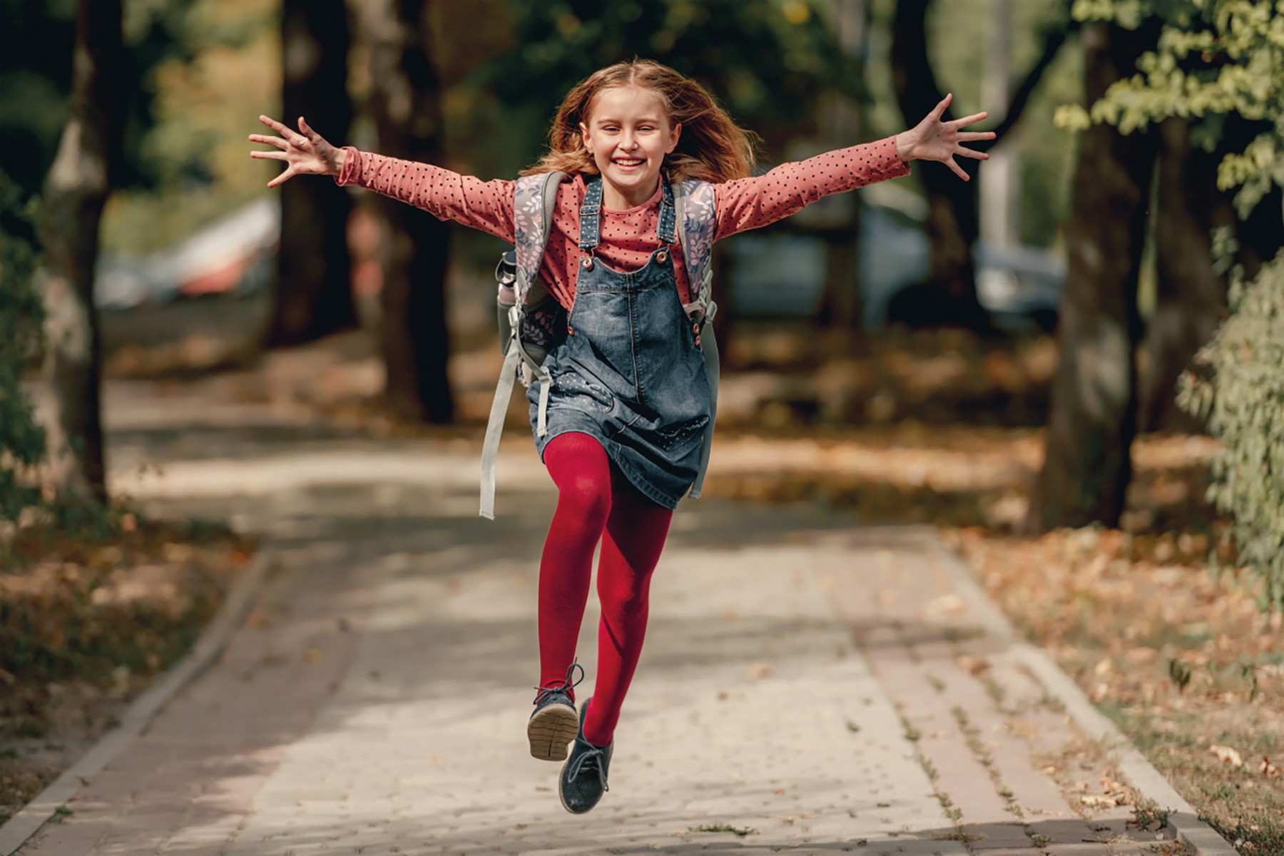 Ein Mädchen mit Schulrucksack springt mit ausgestreckten Armen einen Gehweg entlang.
