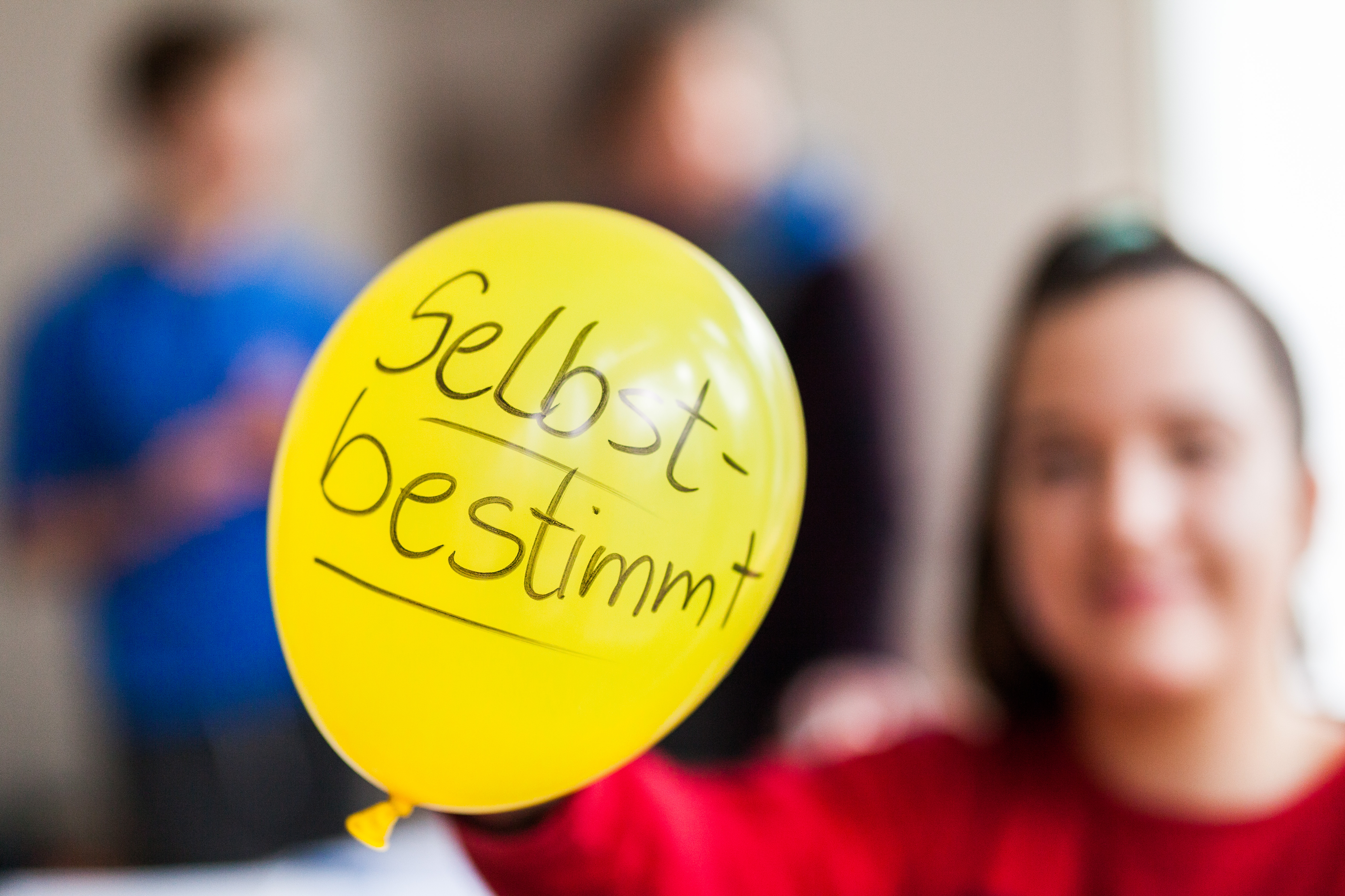 Zu sehen ist ein gelber Luftballon, auf dem "selbstbestimmt" steht. Im Hintergrund sieht man verschwommen eine junge Person, die den Luftballon hält.