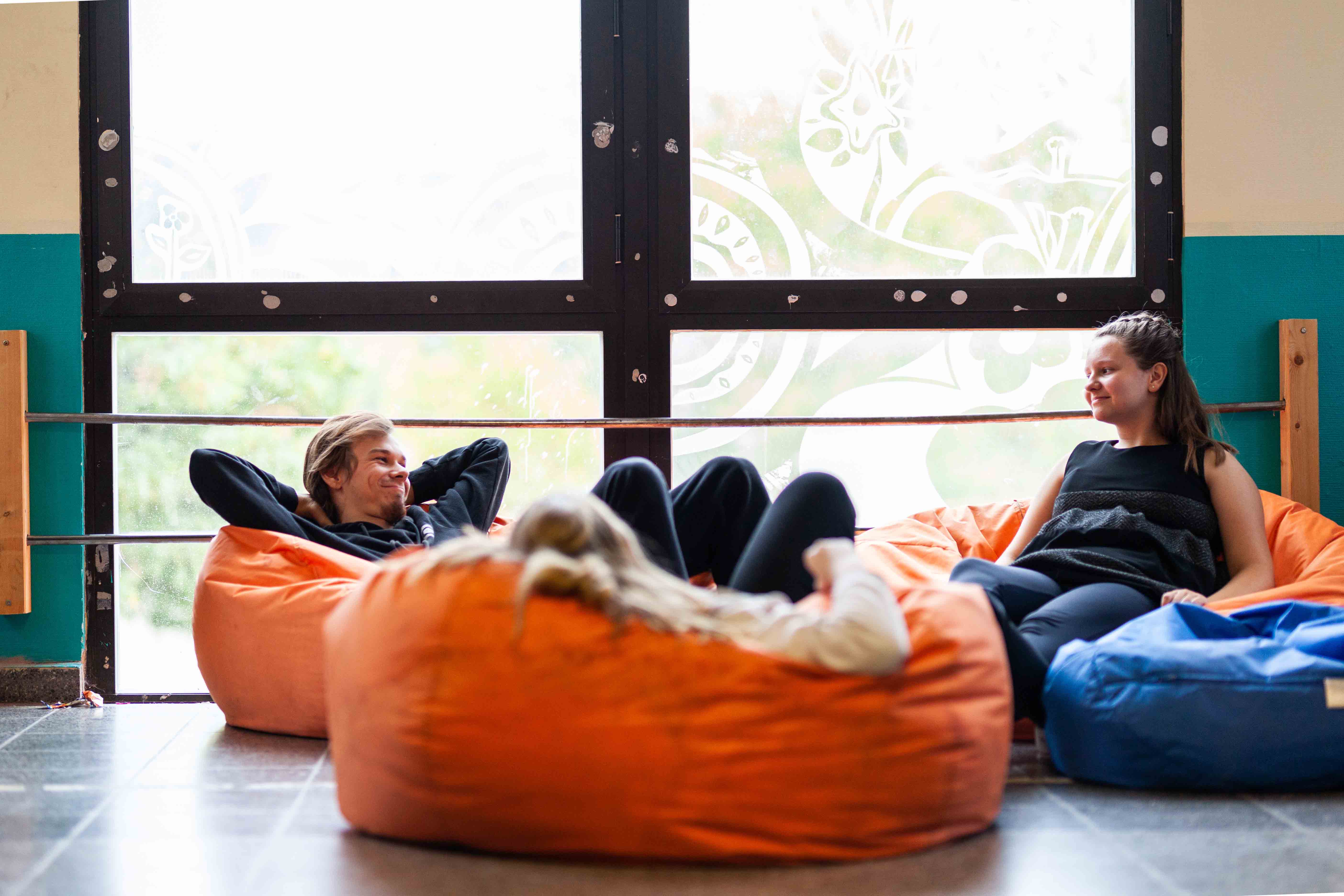 Drei junge Menschen chillen auf orangenem Sitzsäcke vor einem großen Fenster.