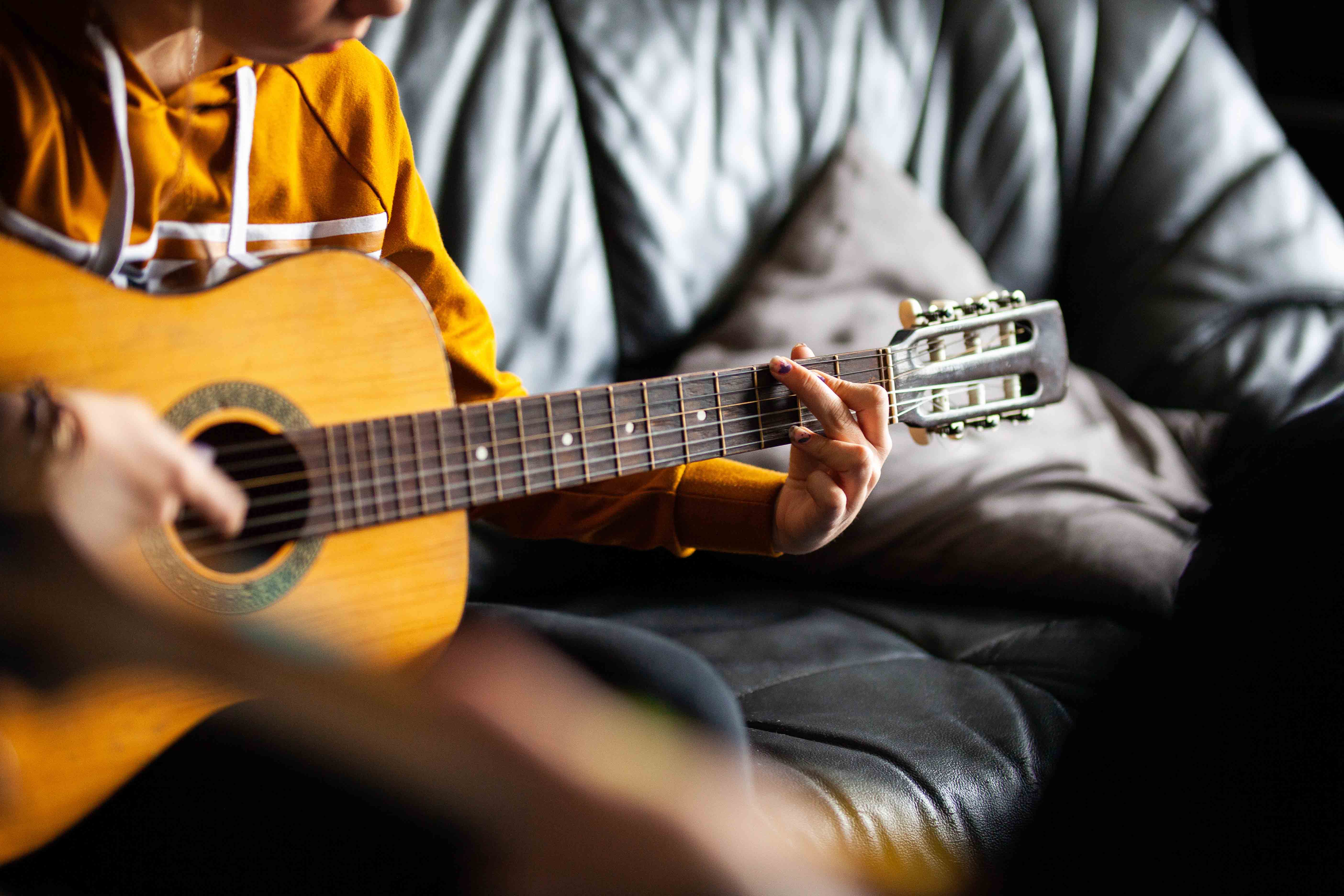 Man sieht im Zentraum eine Gitarre, die von einem Menschen in einem gelben Pullover gespielt wird, die auf einer schwarzen Couch sitzt.