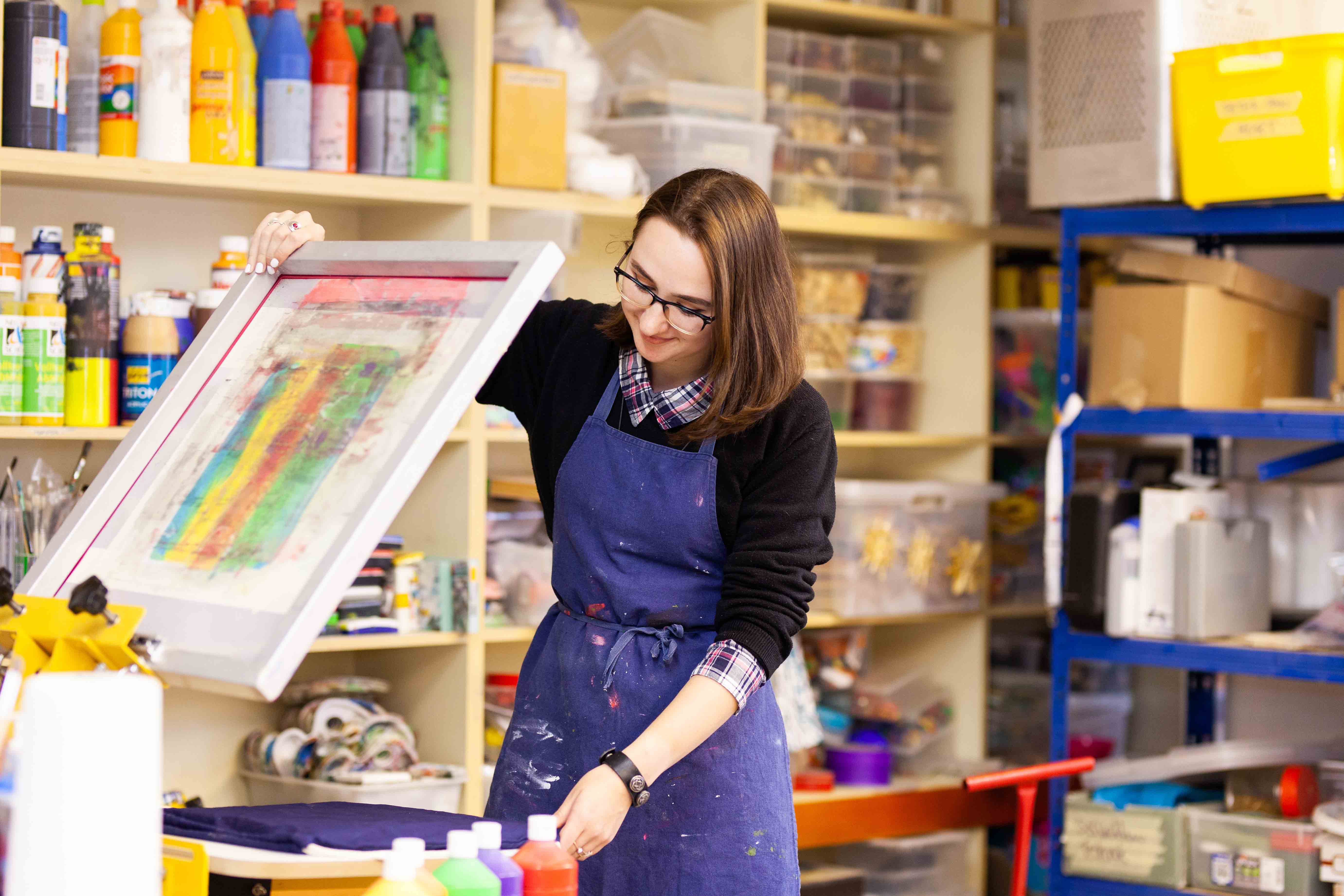 Man sieht eine junge Frau in einer blauen Schürze an einer Siebdruckmaschine. Sie steht in einer Art Werkstatt, um sie herum sind Regale mit bunten Farben und verschiedenen Materialien.