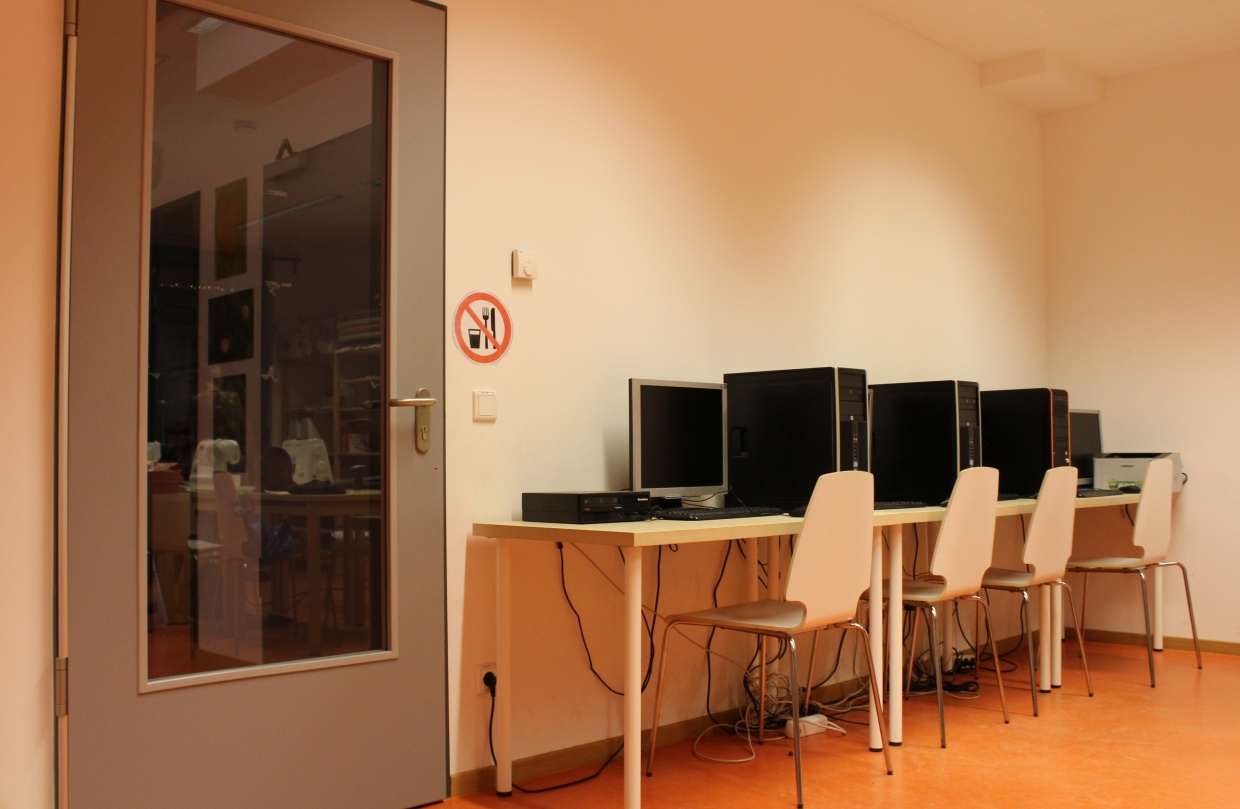 Links ist eine große graue Tür. Rechts davon stehen drei Tische mit jeweils einem Computer und Stühlen davor.
