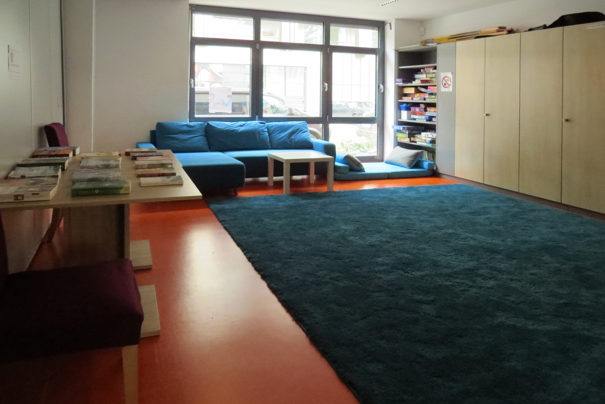 In einem großen Raum liegt ein großer blauer Teppich in der Mitte. Am hinteren Ende steht eine große blaue Couch, an den Wänden links steht ein Tisch mit Büchern, rechts stehen Schränke. In einem davon lassen sich viele Spiele erkennen.