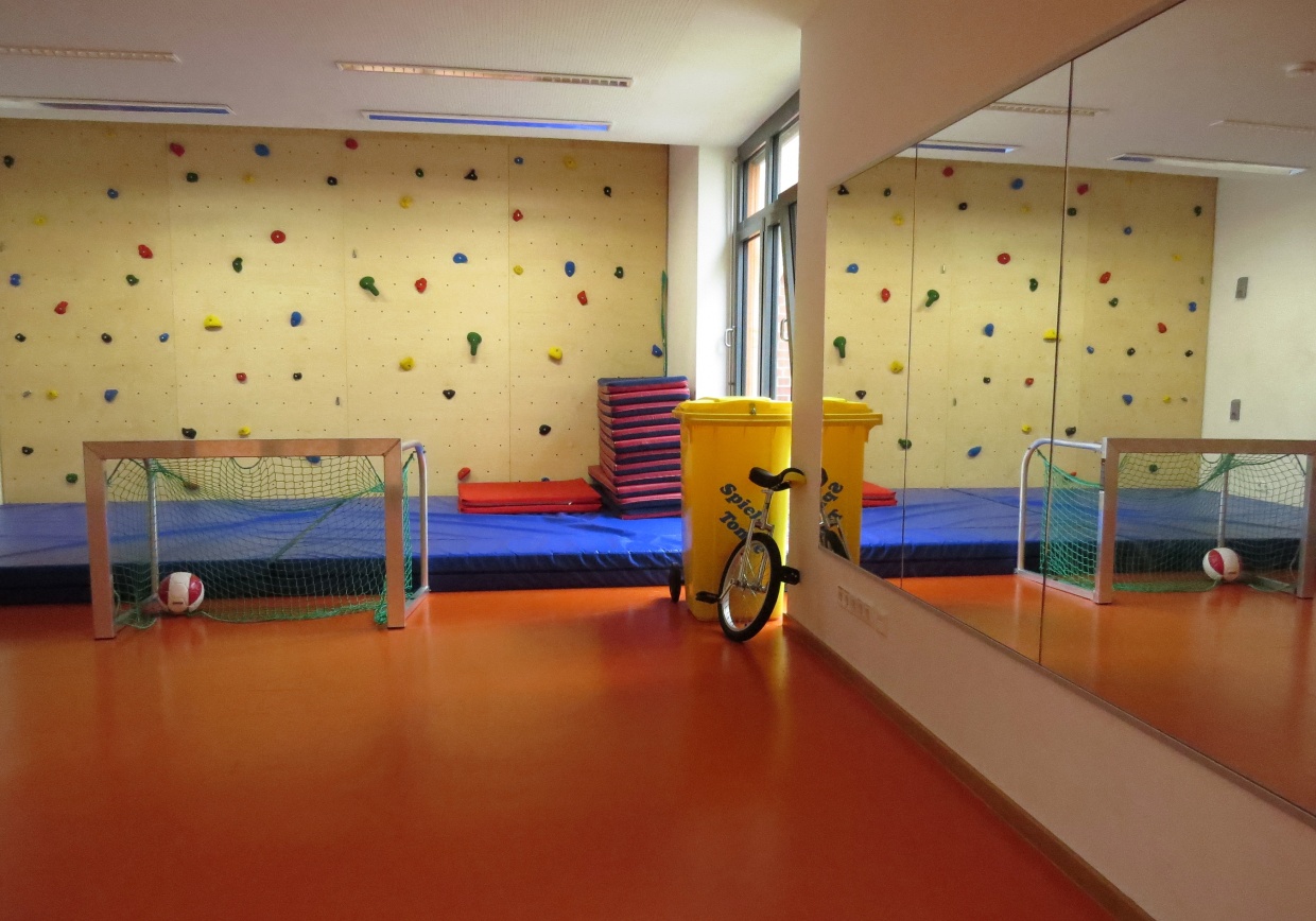 Man sieht eine Ecke von einem großen Sportraum. Rechts an der Wand sind große Spiegel befestigt. An der hinteren Wand erkennt man eine Bowlderwand mit großen Matten. Davor steht ein Hockeytor.
