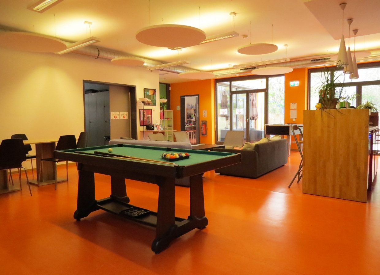 Zu sehen ist ein großer heller Raum mit orangenem Boden. Im vorderen Bereich steht ein Billardtisch, hinten sieht, Couches und Stühle, einen Computerbereich und eine Theke.