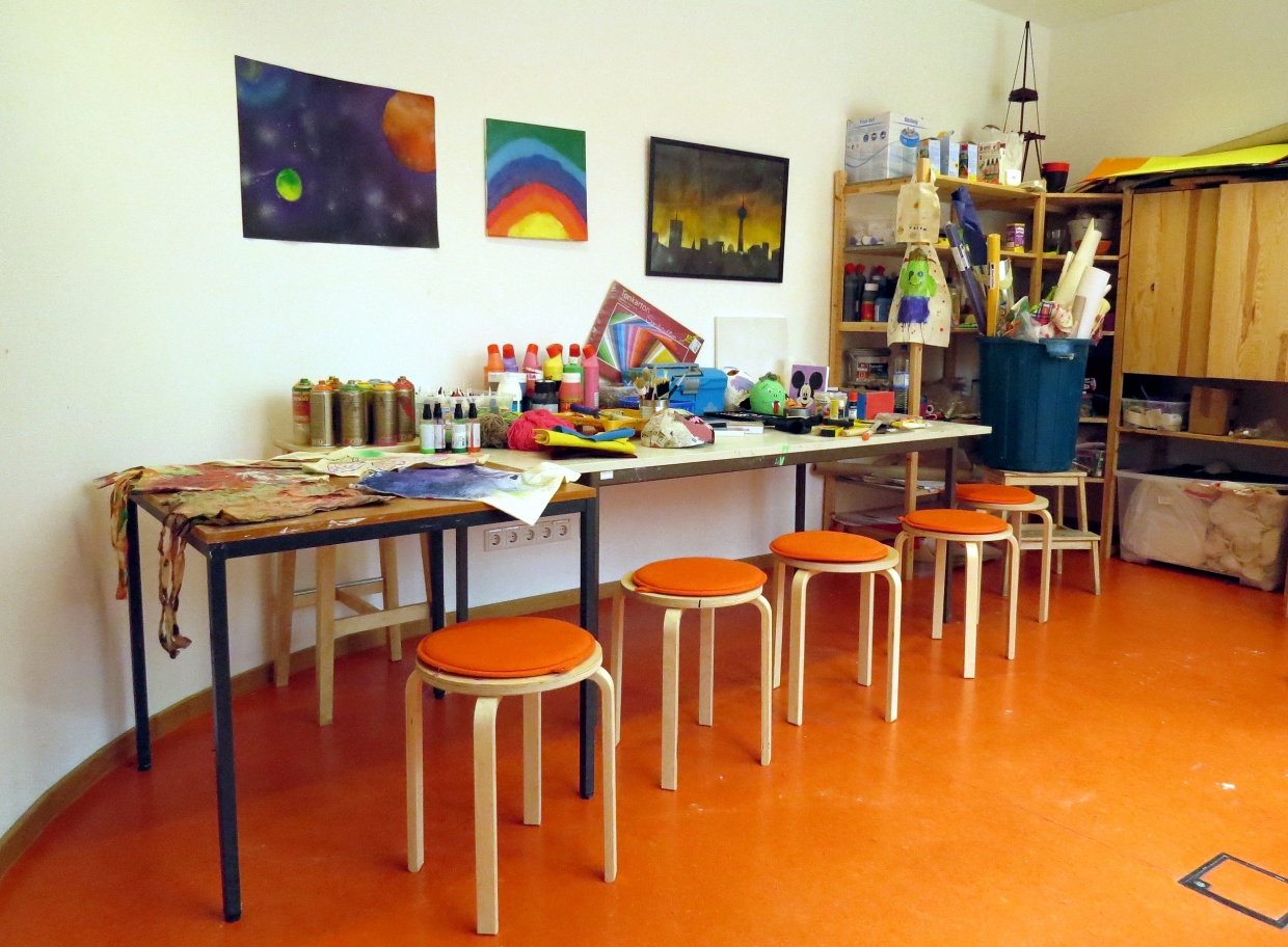 An einer runden Wand stehen mehrere Tische aneinander gestellt, davor jeweils einige Hocker. Auf den Tischen liegt buntes Mal- und Bastelmaterial und darüber hängen selbstgemalte Bilder. Rechts davon steht ein Holzschrank mit noch mehr Farben und Materialien.