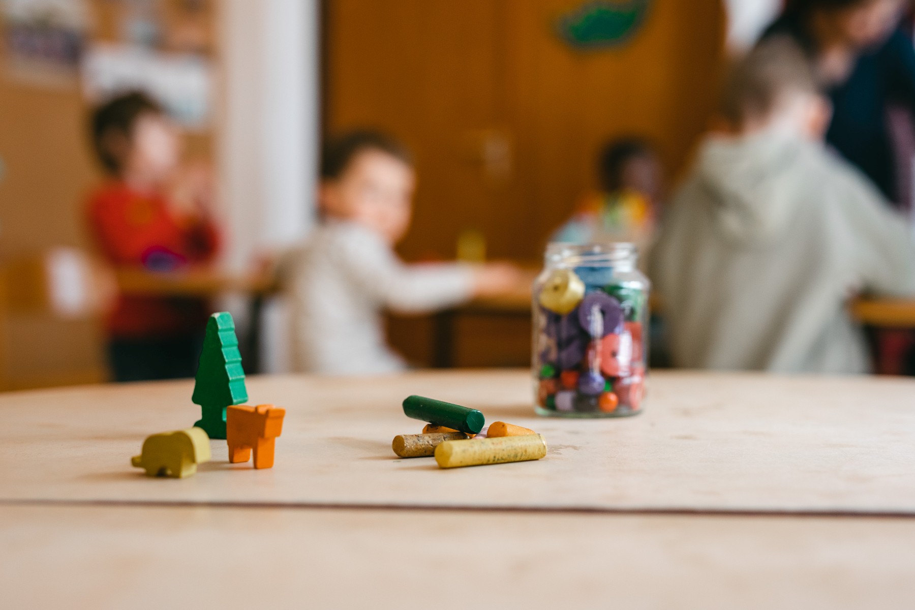 Auf einem Tisch stehen lHolzfiguren und ein Glas mit Fettstiften, einige Stifte liegen vor dem Glas. Im Hintergrund sind unscharf fünf Kinder zu sehen.