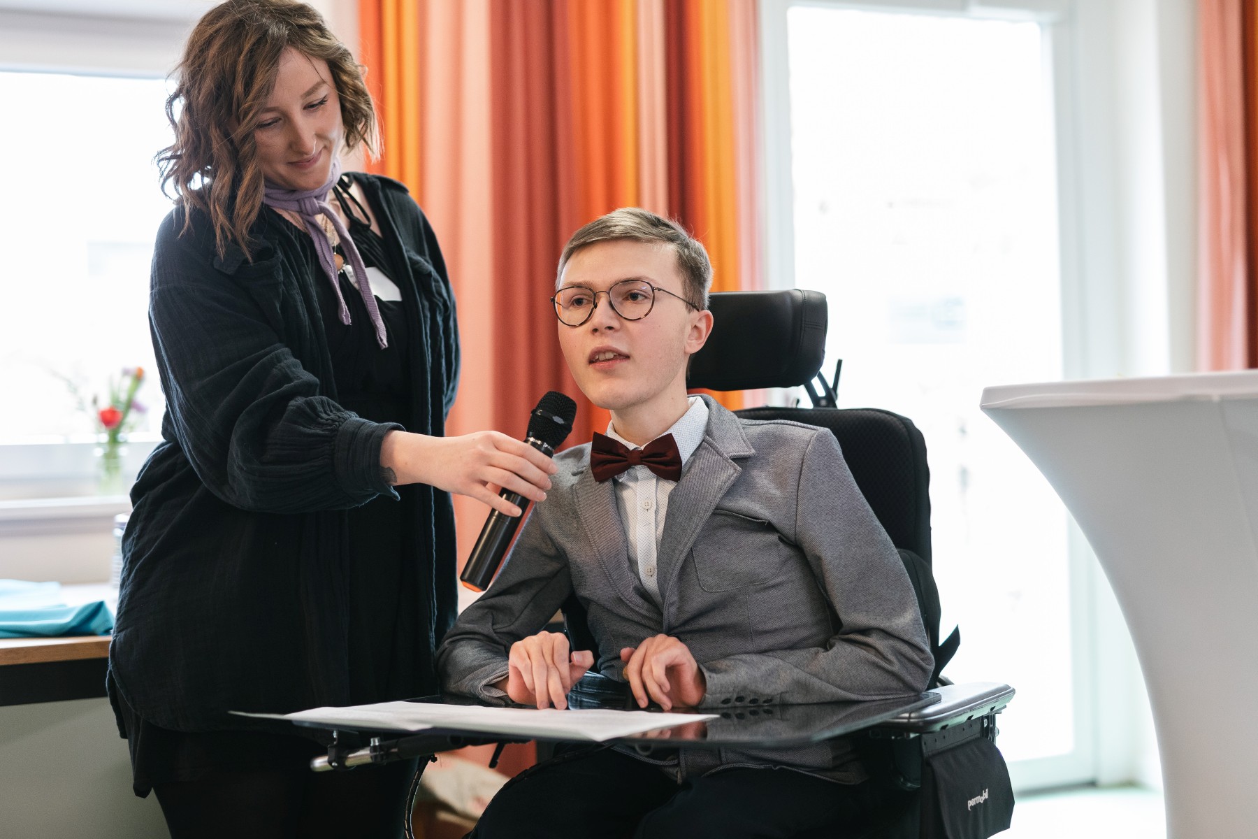Lorenz Klar sitzt im Rollstuhl. Links daneben steht eine Frau, die mit ihrer rechten Hand ein Mikrofon vor seinen Mund hält, in das er spricht.