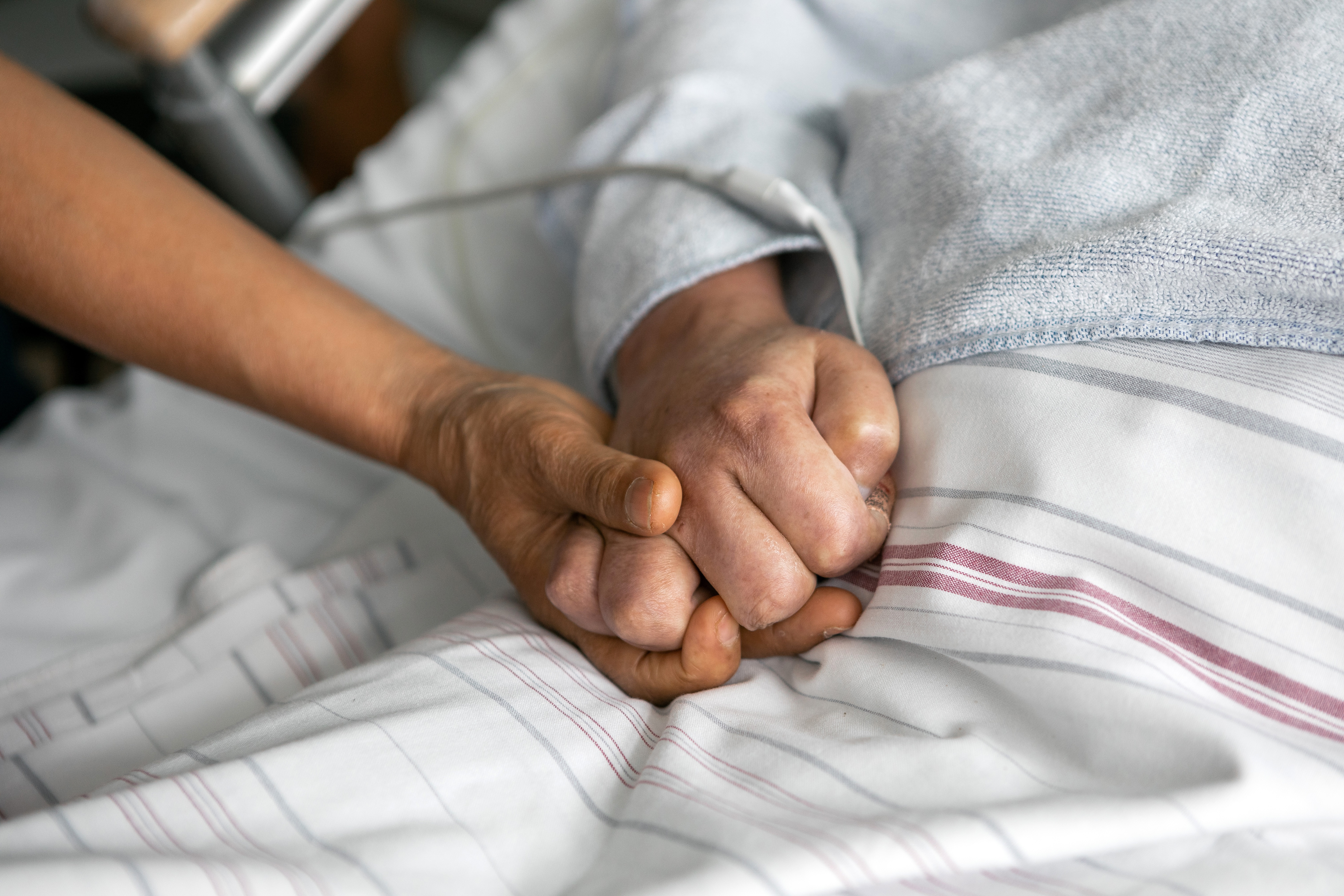 Zu sehen sind zwei Hände, die sich drücken. Eine Hand gehört zu einer Person, die in einem Bett liegt und in der ein Schlauch steckt.