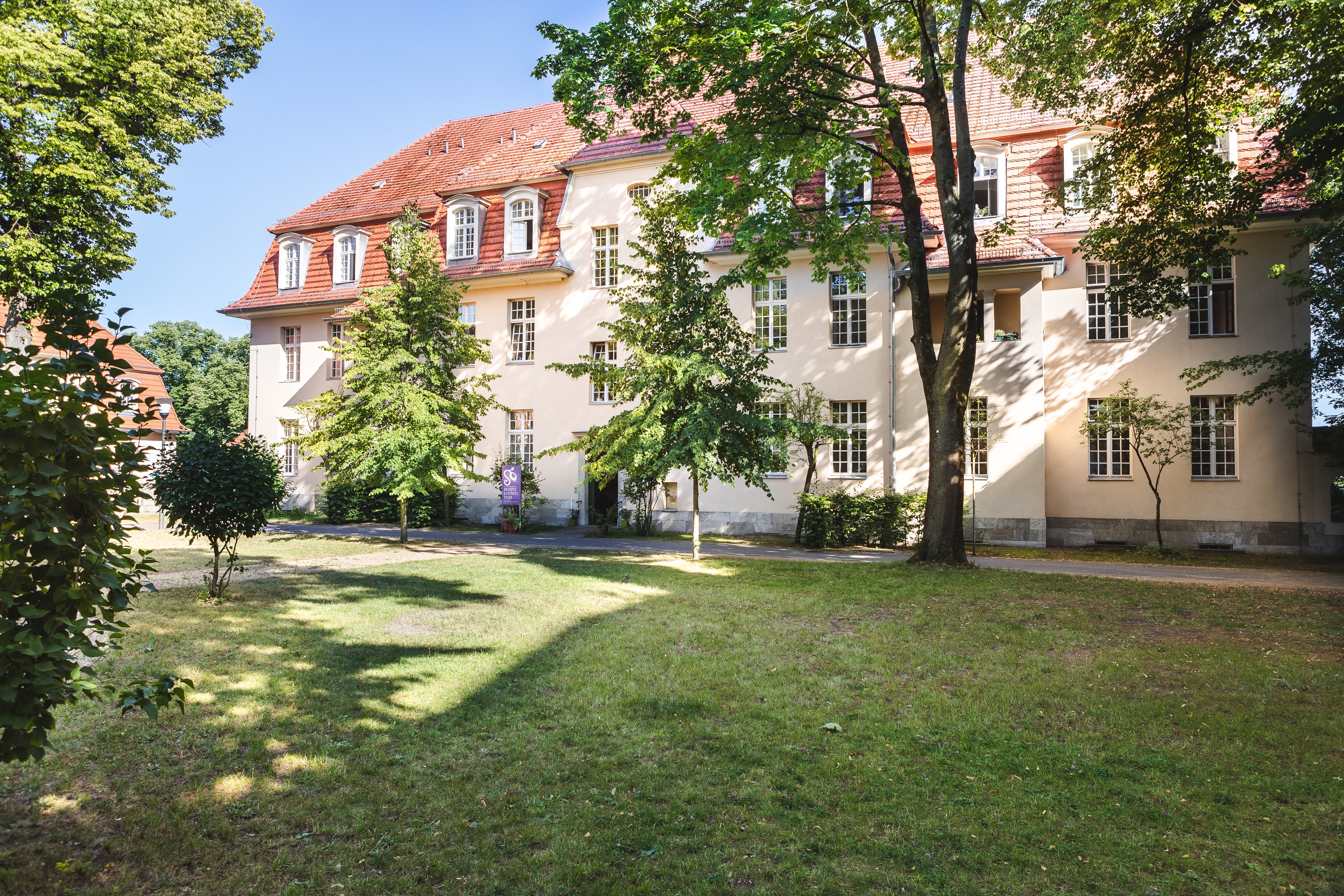 Zu sehen ist eine große grüne Wiese mit Bäumen. Hinten steht das Hospiz LudwigPark: ein großes helles und breites Haus mit vielen Fenstern.