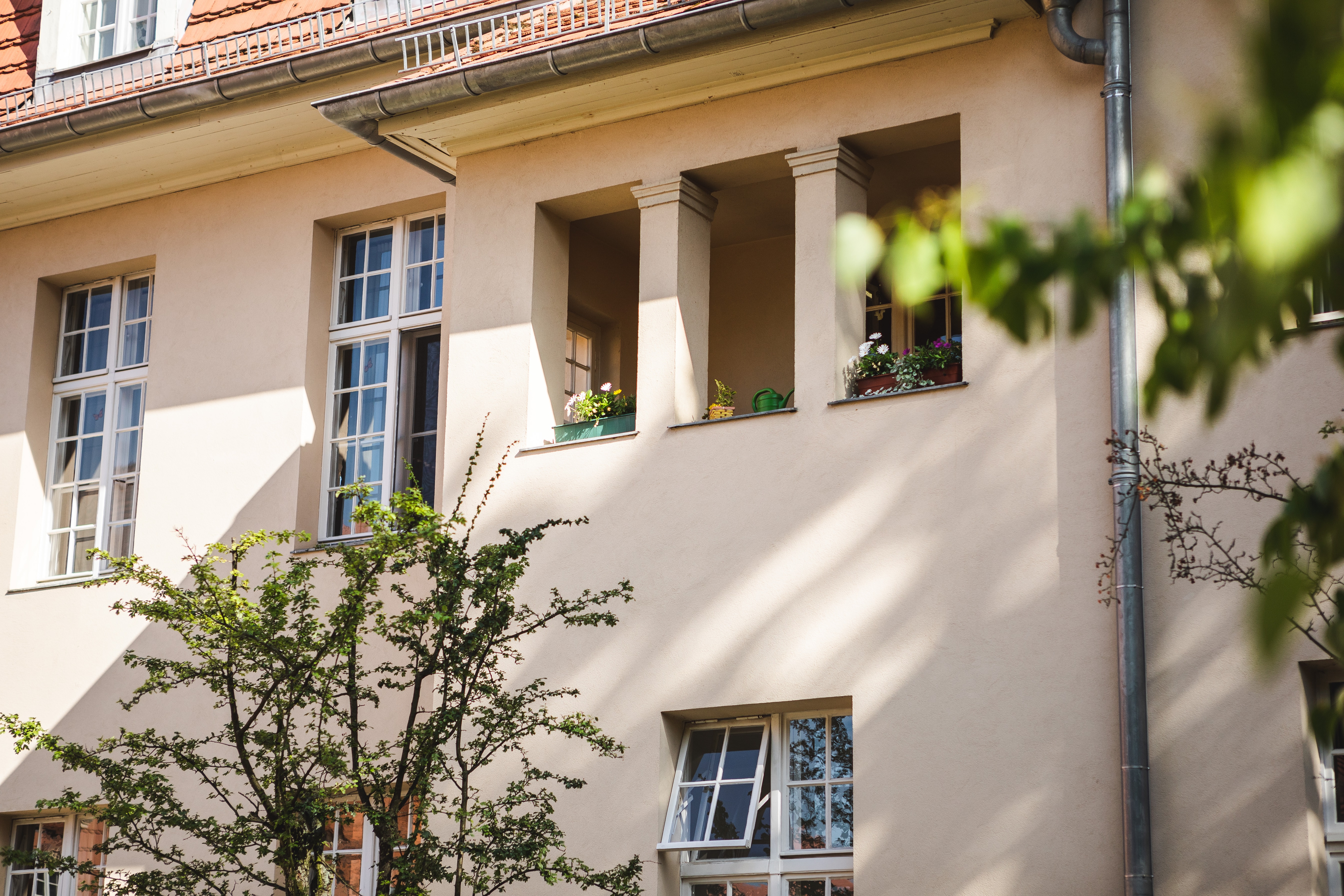 Zu sehen ist eine helle Hauswand mit vielen Fenstern. Es ist Sommer, favor stehen ein paar Bäume.