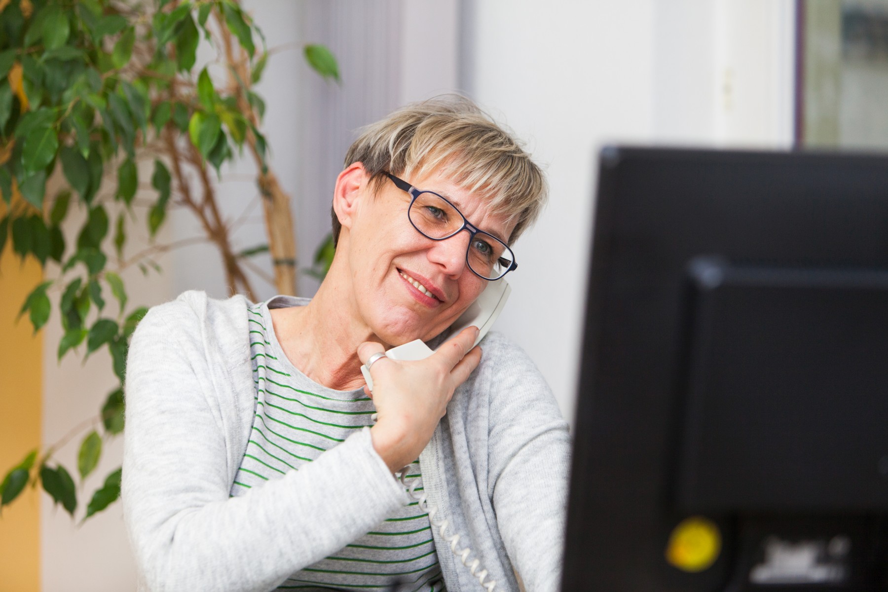Zu sehen ist eine Frau mit einer Brille und kurzen, blonden Haaren, die mit ihrer Schulter ein weißes Telefon an ihr Ohr drückt und auf einen Computerbildschirm vor sich schaut. Sie lächelt leicht.