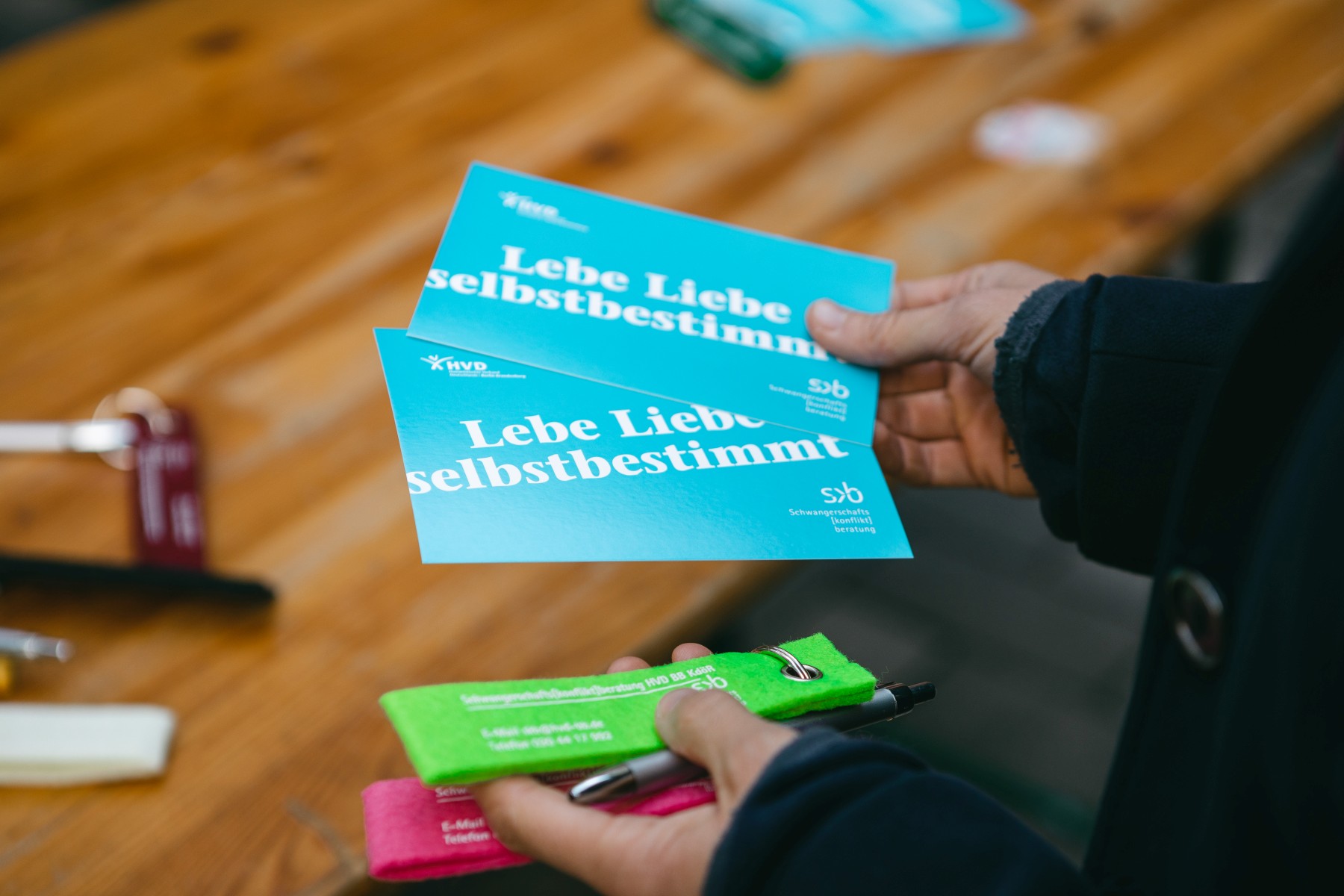 Die rechte Hand einer Person hält zwei Flyer der Schwangerschafts[konfikt]beratung in Berlin-Pankow, in der linken Hand liegen ein silbergrauer Kugelschreiber und zwei Schlüsselanhänger der Einrichtung: einer in Hellgrün, einer in Pink.