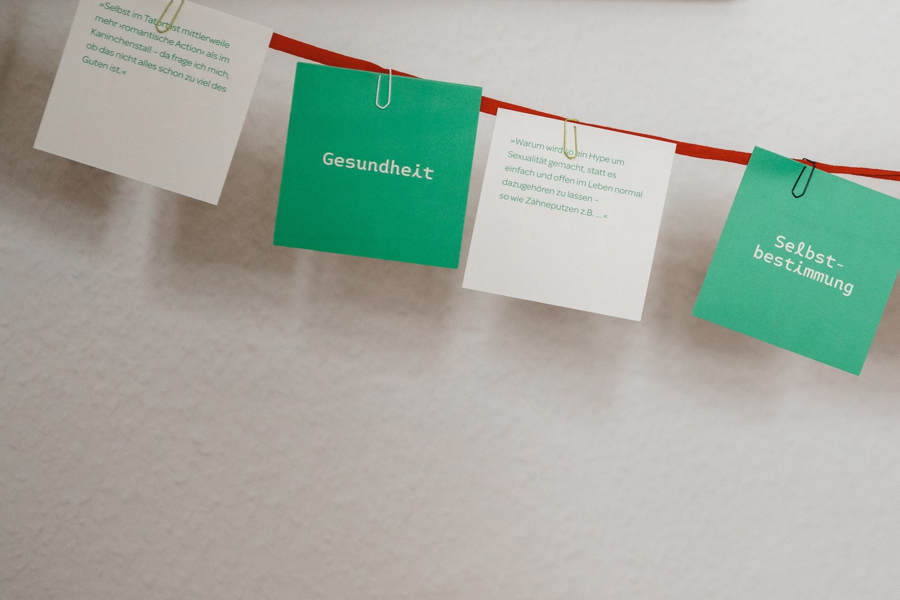 Vor einer weißen Wand hängen an einer roten Schnur abwechselnd, mit je einer Büroklammer befestigt, ein weißer, dunkelgrüner, weißer und dunkelgrüner quadratischer Zettel. Auf den weißen sind Zitate von Personen, die beraten wurden, zu lesen und auf den grünen einmal „Gesundheit“ und einmal „Selbstbestimmung“.