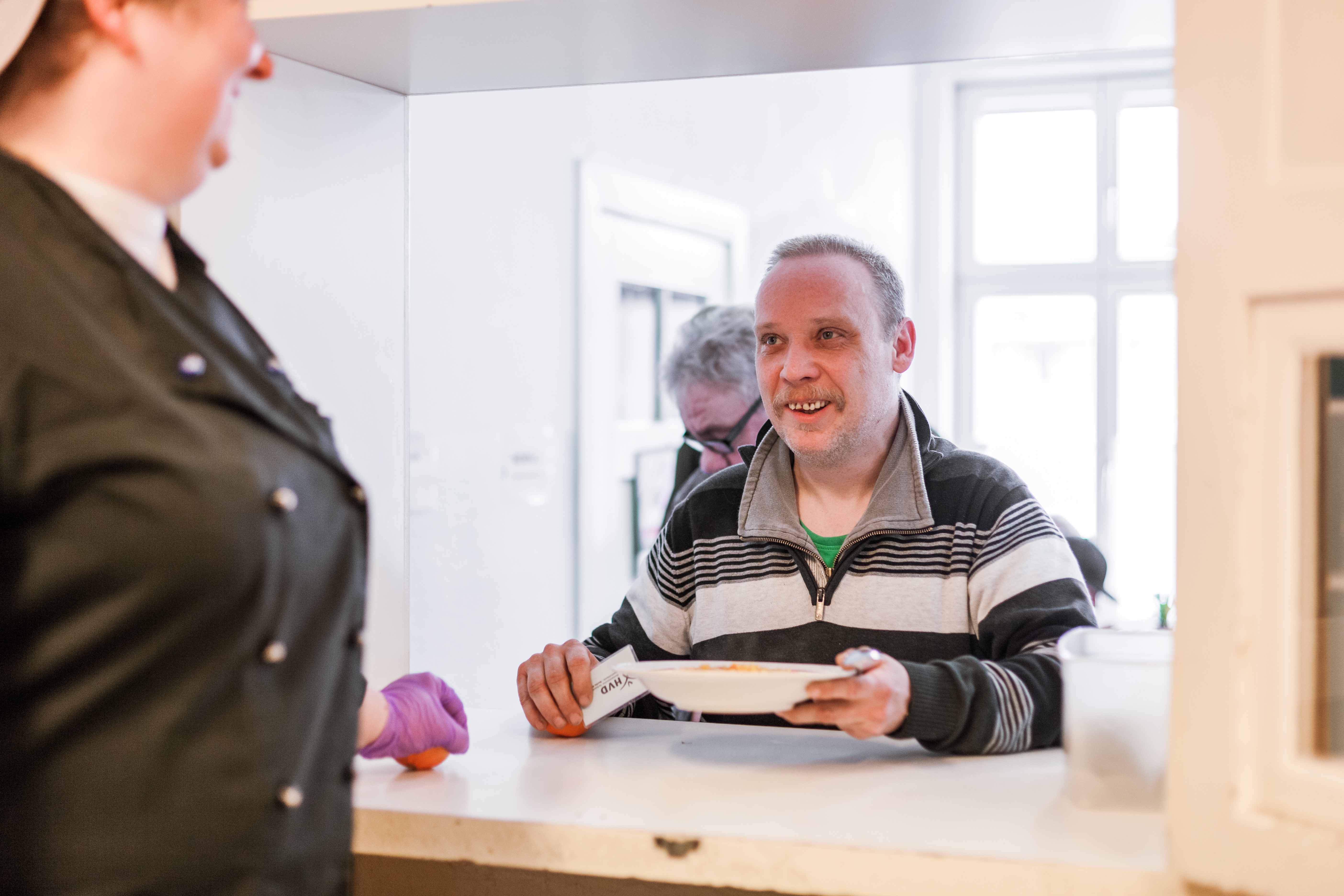 Links steht angereissen eine Frau in einer schwarzen Kochjacke und schaut druch die Luke der Essensausgabe auf einem Mann, der sich gerade einen Teller mit warmen Essen und eine Mandarine von der Theke nimmt. Sie lächeln sich an. 