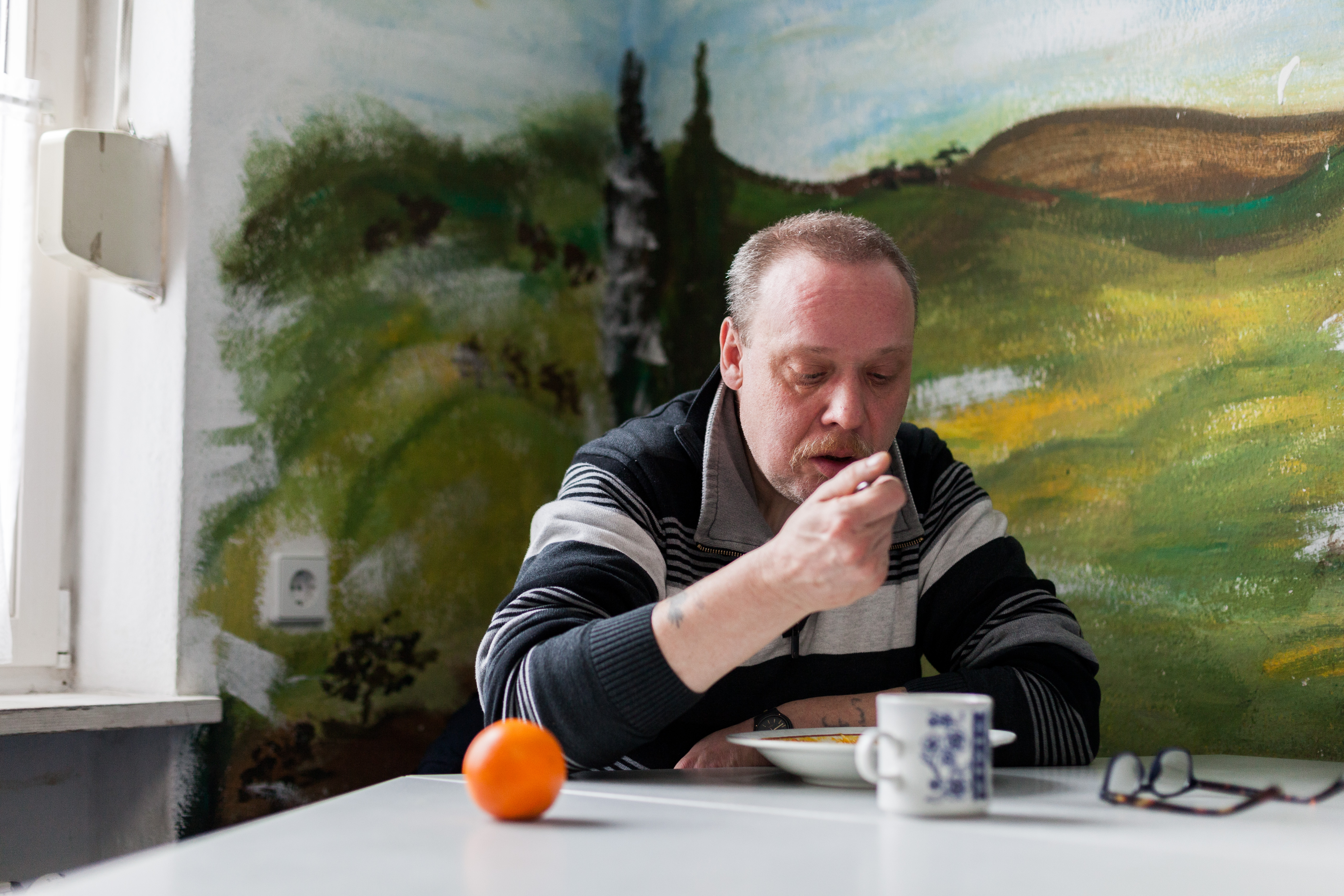 Ein Mann sitzt in einer Ecke an einem Tisch und isst gerade von einem Löffel. Vor ihm steht ein gefüllter Teller, eine Tasse, eine Brille und eine Mandarine.