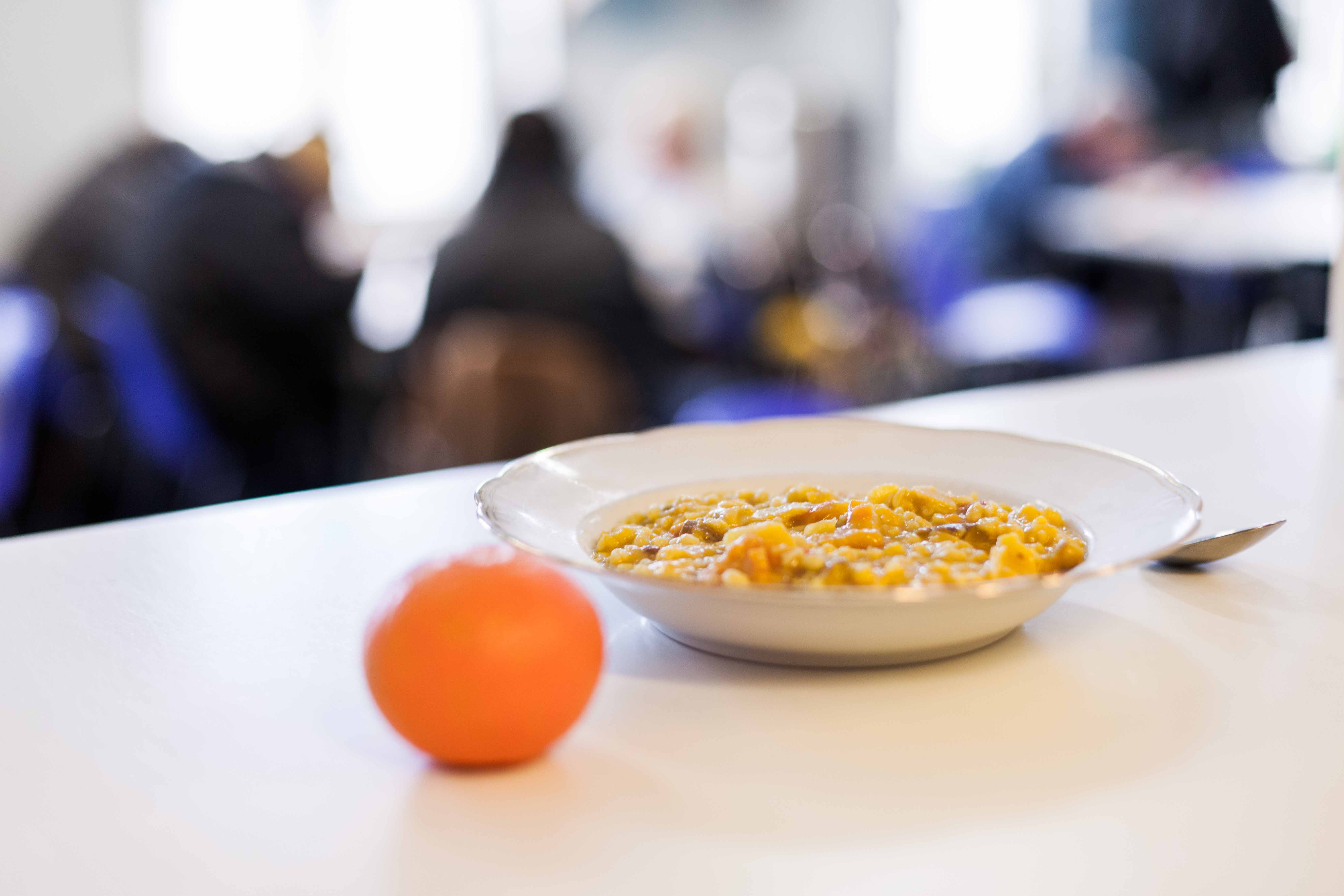 Auf einem weißen Tisch steht ein mit warmen Essen gefüllter Teller. Daneben liegt eine Mandarine und ein Löffel.