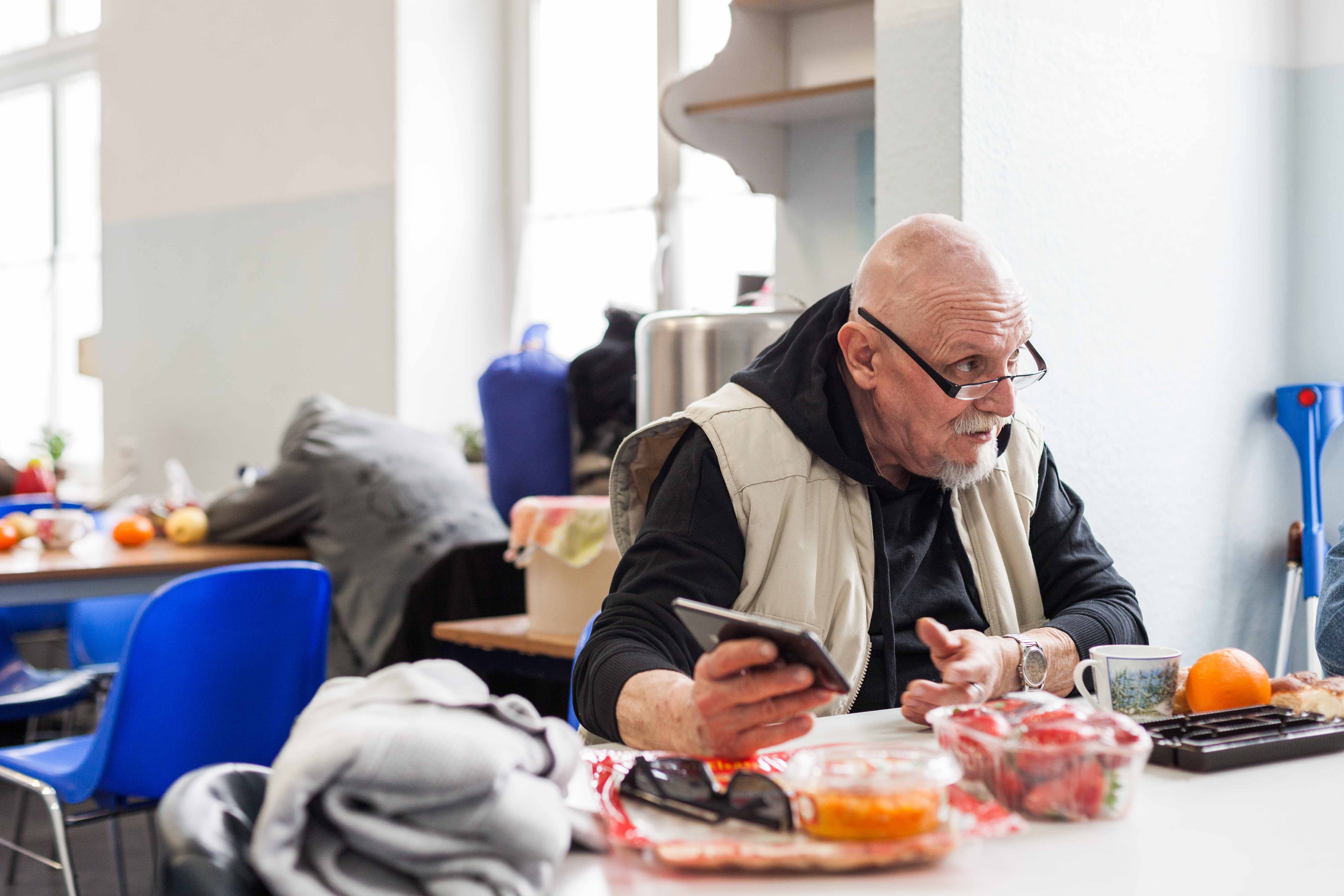Ein Mann in einer beigen Weste und mit einer Brille sitzt an einem Tisch. Vor ihm steht Essen. In der rechten Hand hält er ein Handy und er scheint mit jemanden zu reden. Im Hintergrund sieht man einen weiteren Tisch und verschwommen eine Person, die daran sitzt.