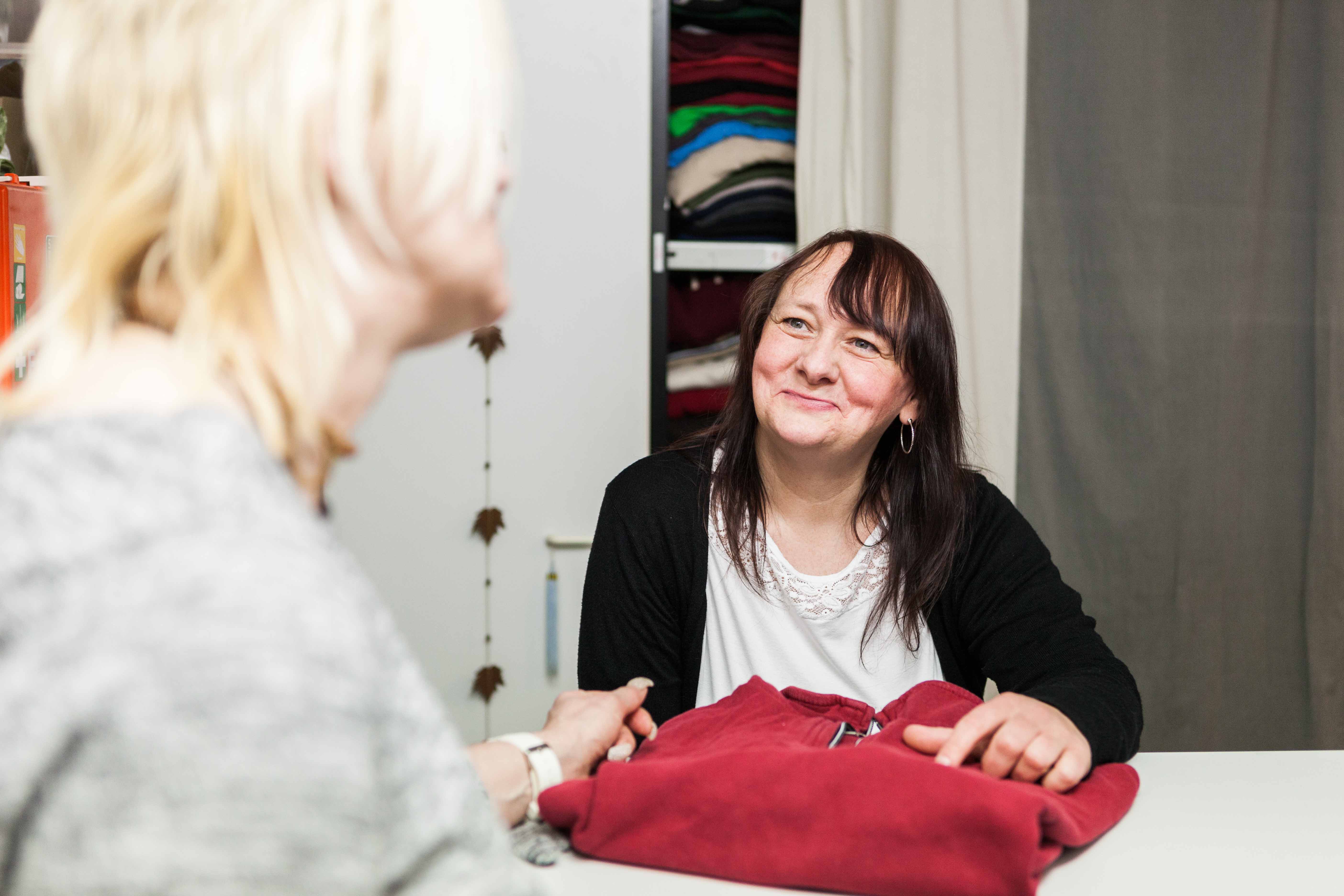 Zu sehen ist eine Frau in einer schwarzen Jacke, die sich über eine Art Theke lehnt. Vor ihr liegt ein roter Pullover, den sie gerade an eine andere Frau vor der Theke aushändigt. Hinter ihr ist ein Schrank mit mehr Kleidung zu erkennen.