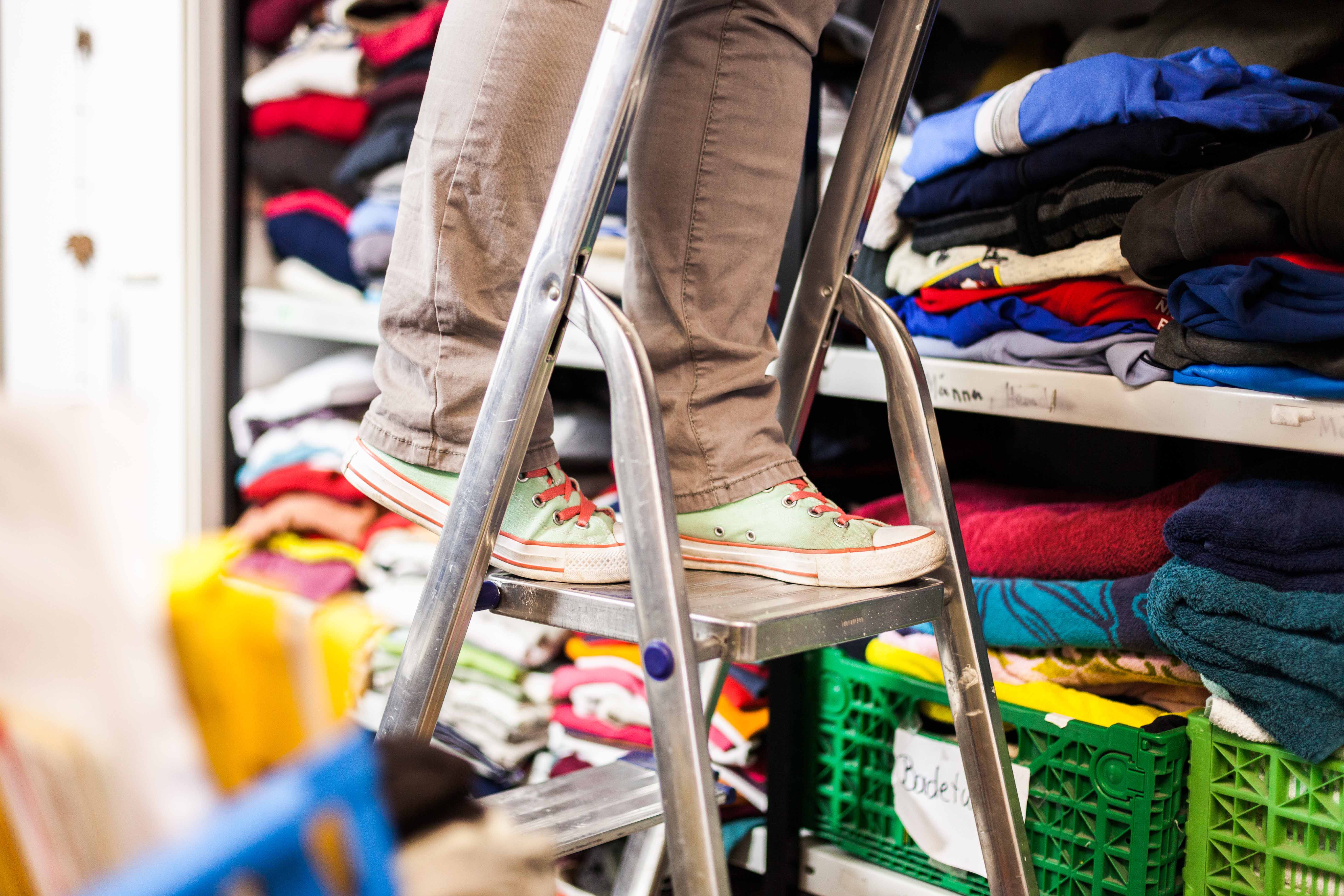 Zu sehen sind die Beine einer Person, die auf einer Leiter steht. Die Leiter steht an einem Schrank, der mit Kleidung gefüllt ist.