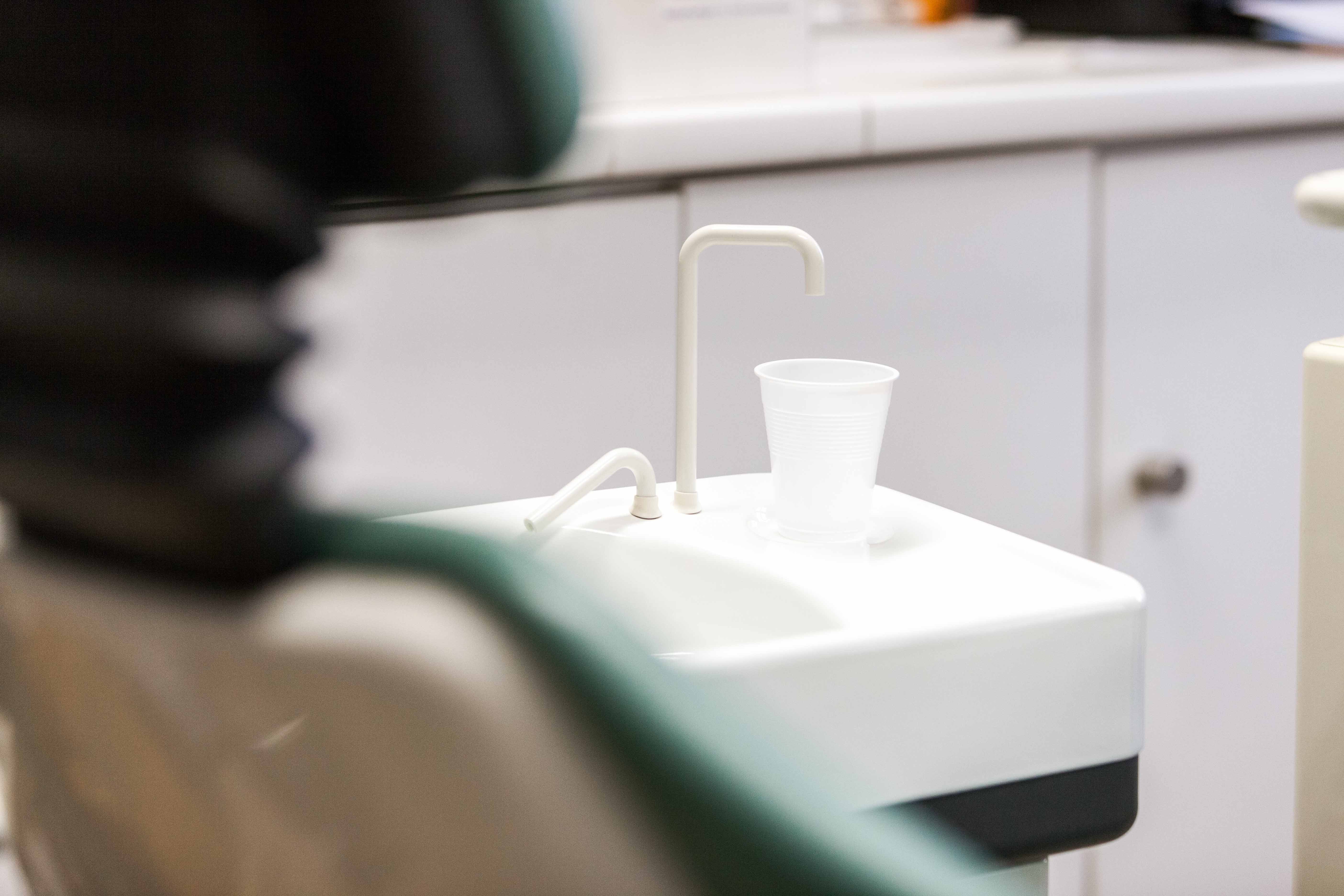 Verschwommen im Vordergrund ist angeschnitten ein Zahnarztstuhl zu erkennen. Im Zentrum sieht man ein kleines Waschbecken mit einem Plastikbecher, wie es in Zahnarztpraxen üblich ist.
