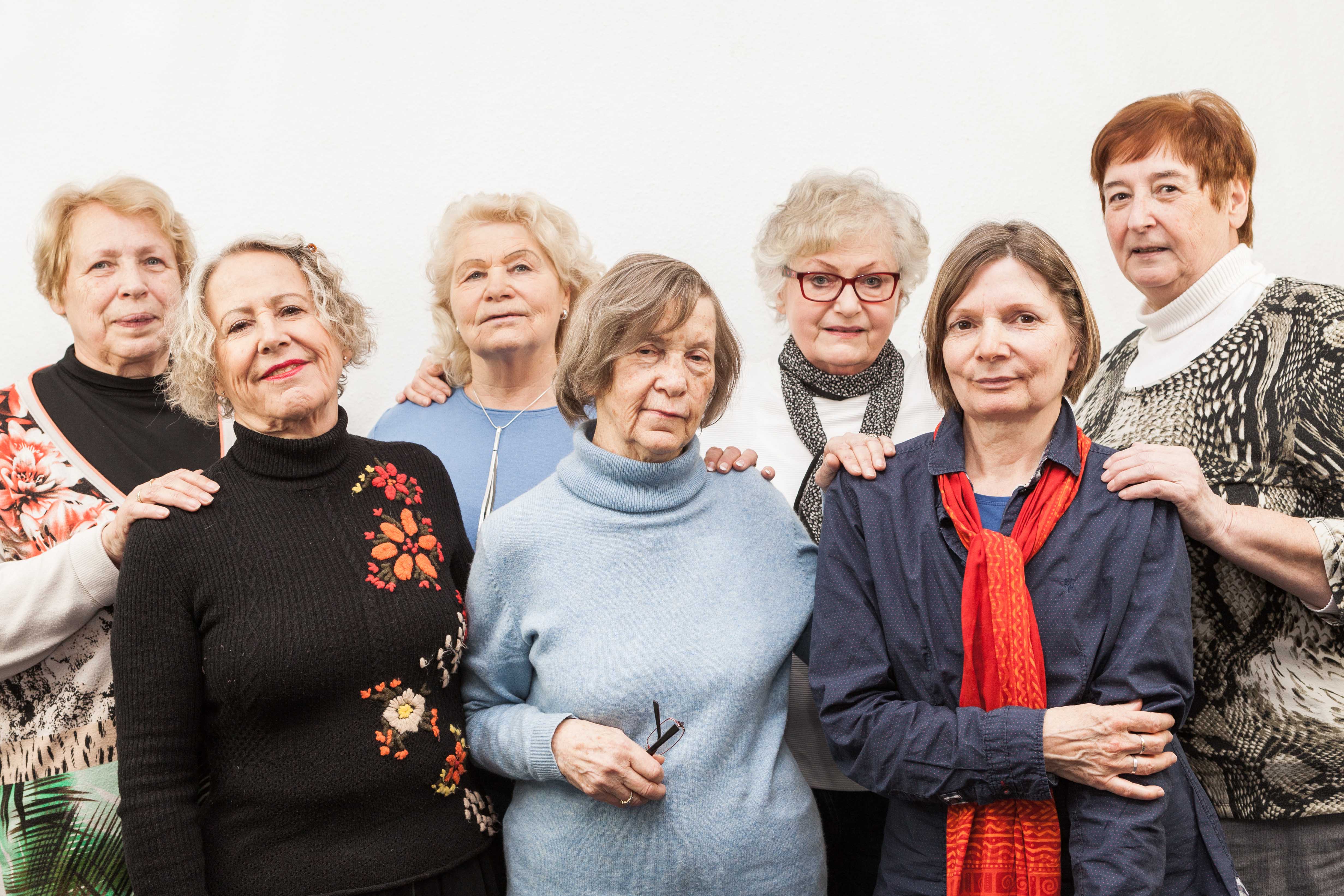 Zu sehen sind sechs ältere Frauen, die in zwei Reihen nebeneinander stehen. Sie schauen in die Kamera und lächeln.