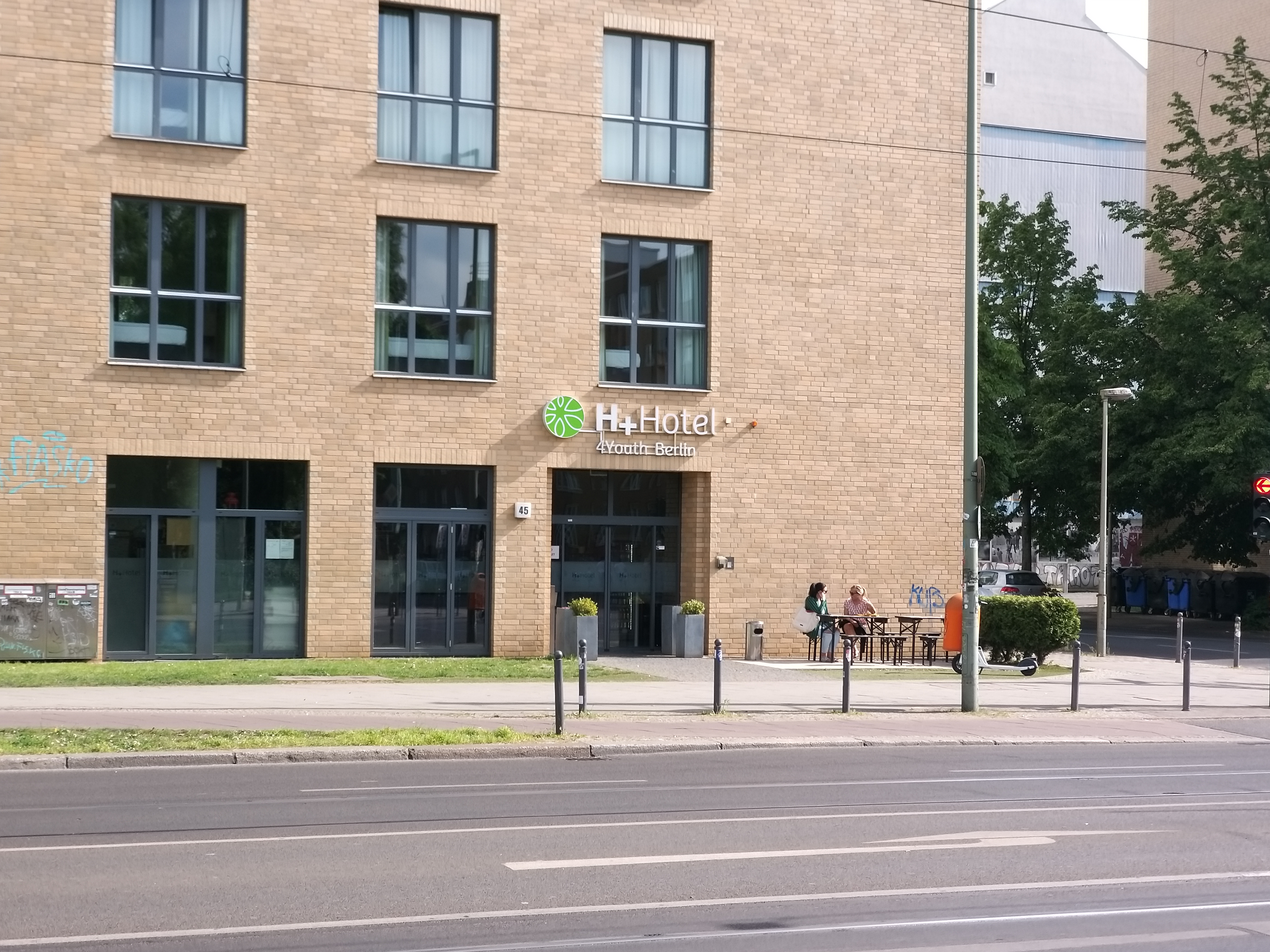 Zu sehen ist eine Hausecke mit großen Fenstern an einer großen Straße. Die Eingagstür ist mit H+ Hotel 4Youth Berlin beschriftet.