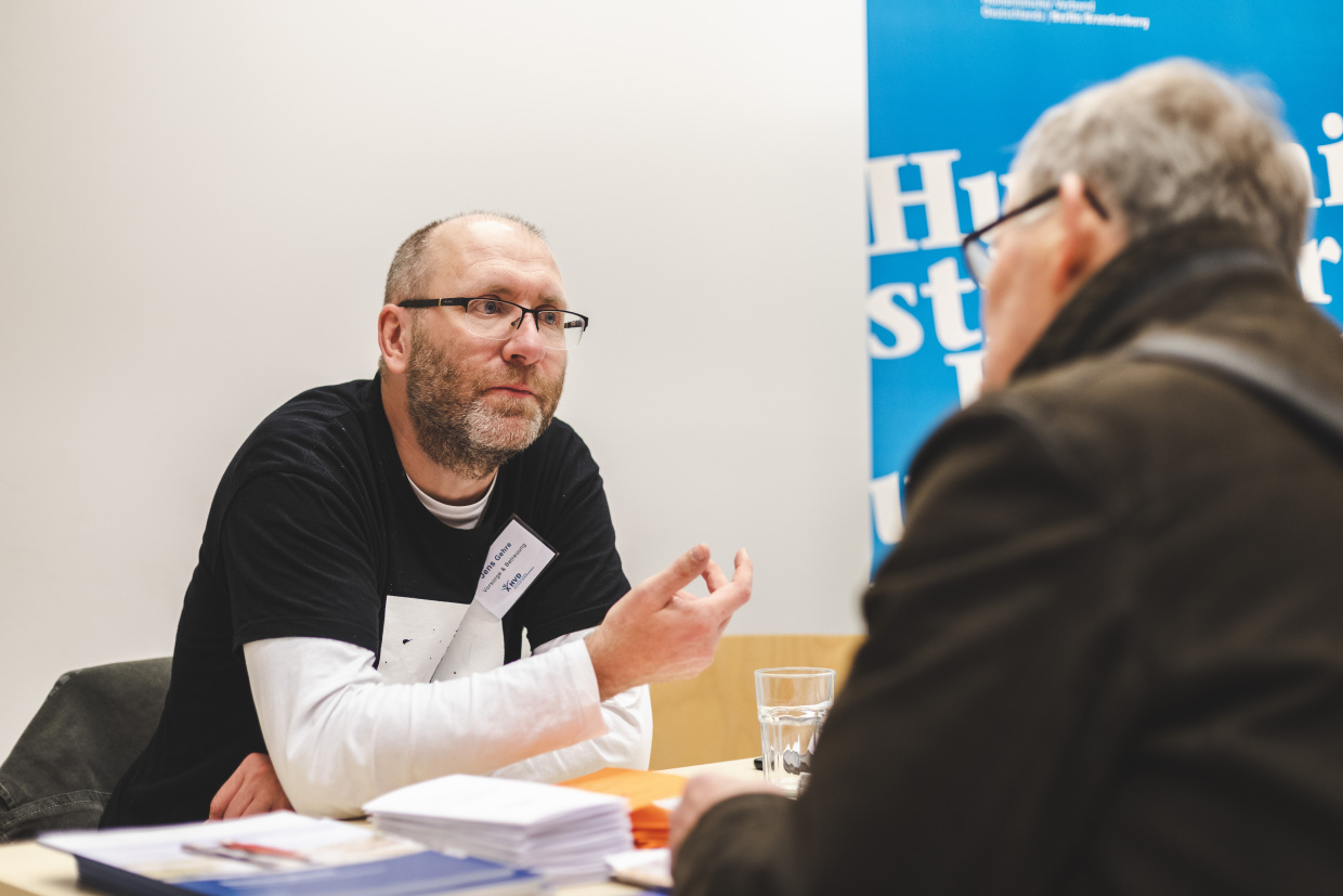 Jens Gehre, Nachlassberater beim Humanistischen Verband Berlin-Brandenburg, sitzt an einem Tisch und berät einen Mann, der ihm gegenüber am Tisch sitzt.