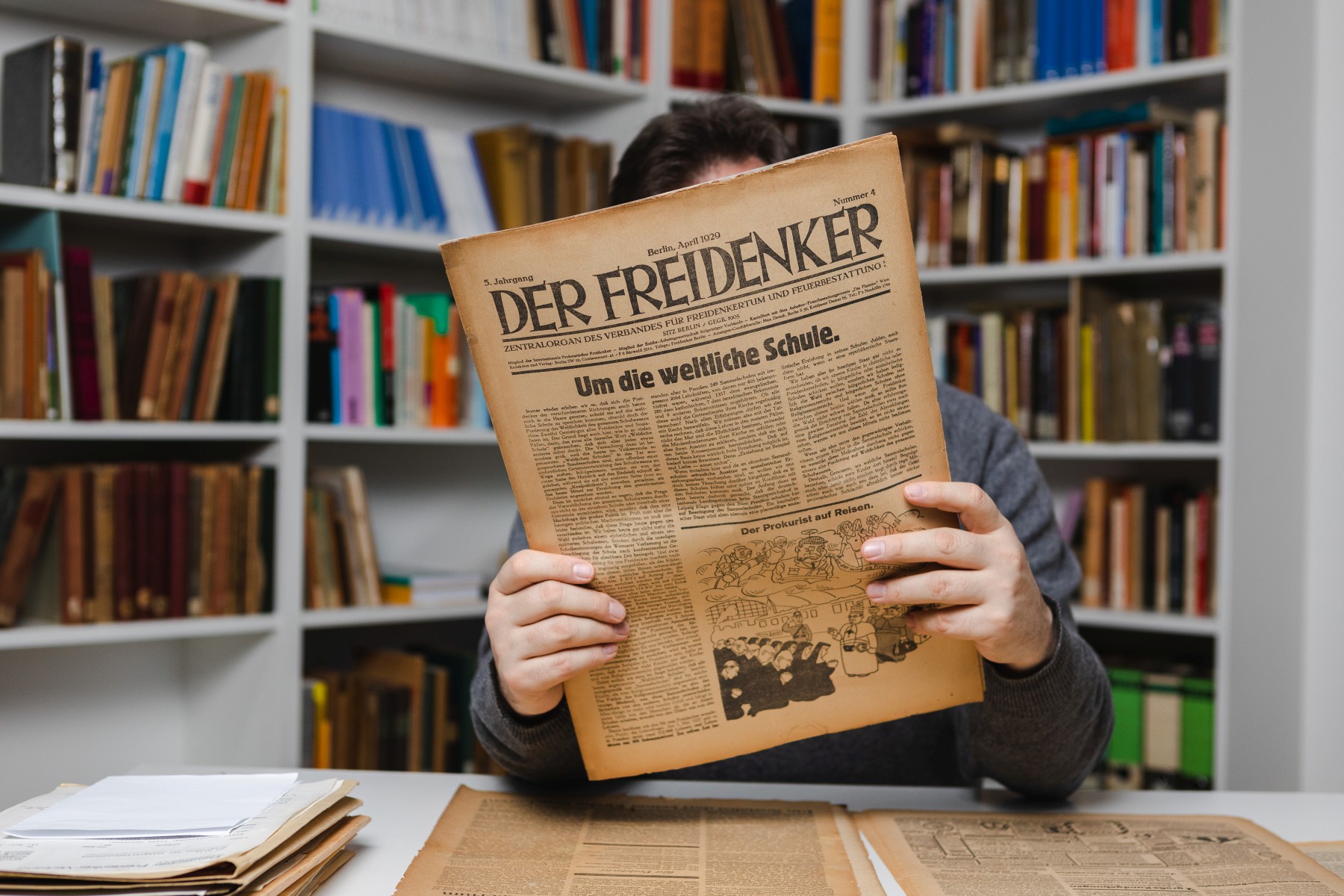 An einem Tisch in einem Raum mit Regalen voller Aktenordner und Bücher sitzt eine Person. Ihr Gesicht wird durch eine historische Zeitschrift verdeckt, die die Person in ihren Händen hält.