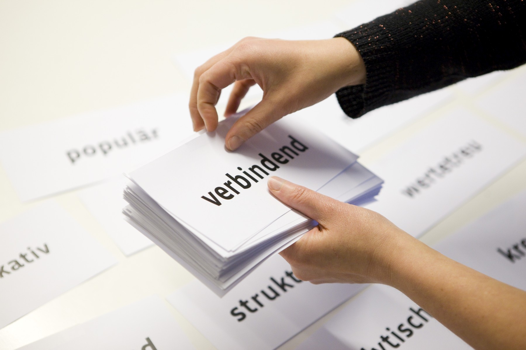Zwei Arme zweier Personen sind zu sehen. Beide Hände halten einen Stapel weißer Zettel. Auf dem oberen Zettel steht in schwarzer Schrift: verbindend.