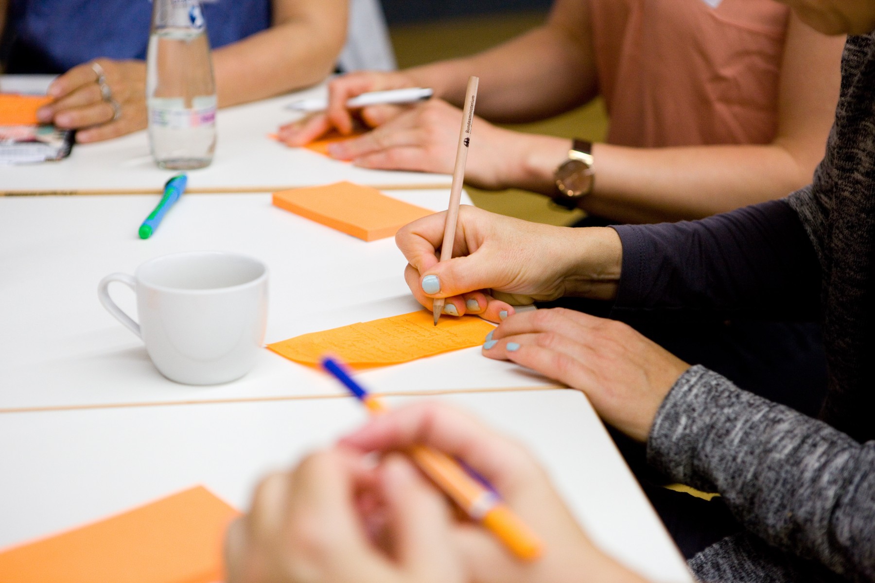 Vier Personen, von den nur die Unterarme oder Hände zu sehen sind, sitzen an einem Tisch. Darauf liege orange Zettel. Ein Person schreibt mit einem Bleistift etwas auf einen solchen Zettel.