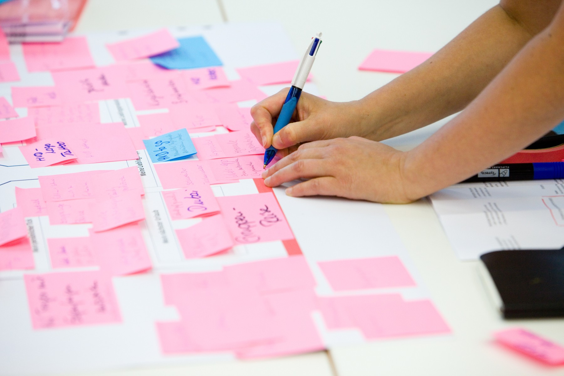 Auf einem Tisch liegen weiße Papierblätter, auf denen beschriebene rosa und blaue Post-its kleben. Eine Person, von der nur die Unterarme zu sehen sind, schreibt mit einem Kugelschreiber etwas auf ein rosa Post-it.