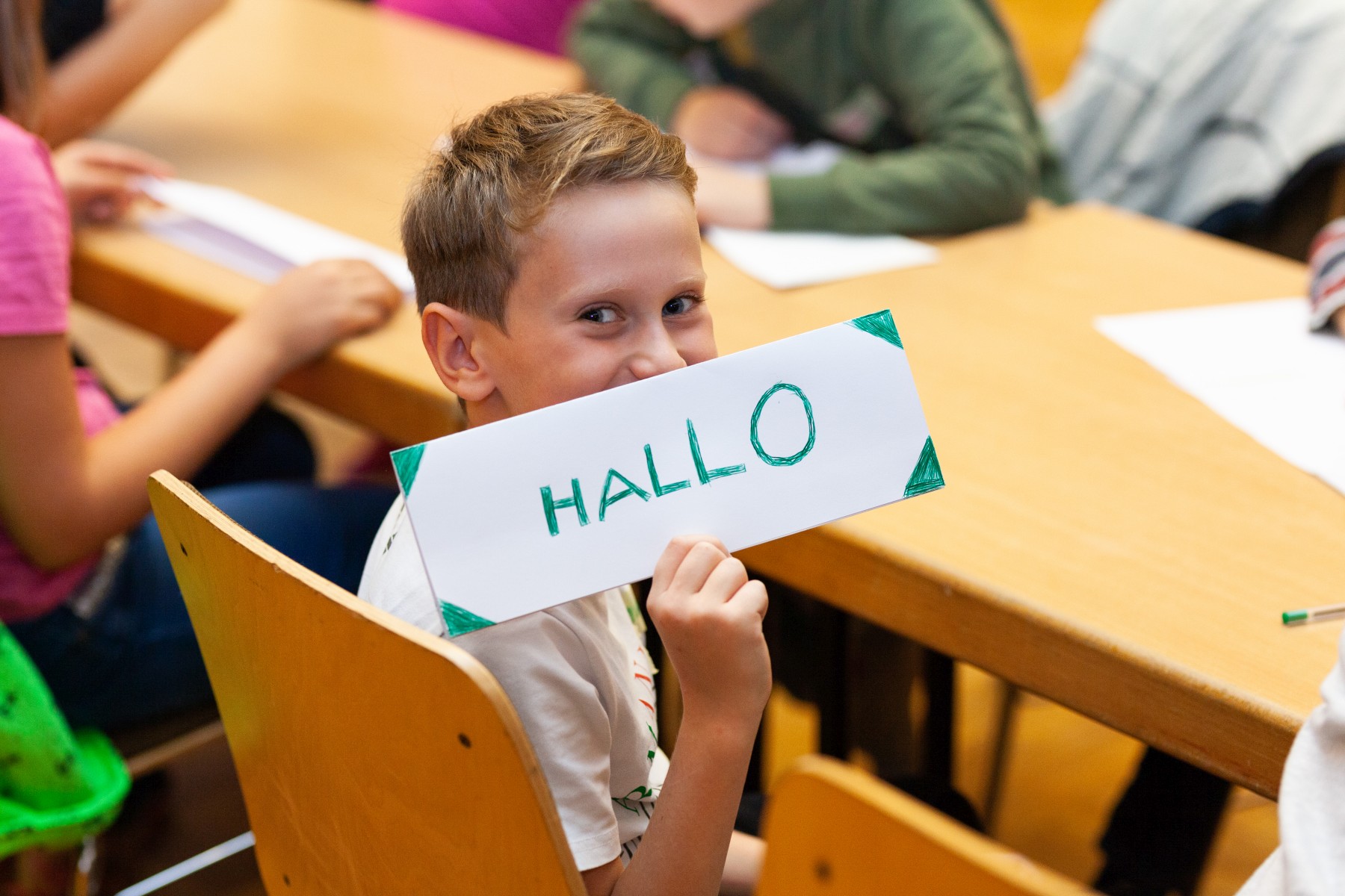 Ein Junge sitzt am Tisch und guckt in Richtung der Person, die dieses Bild hier betrachtet. Er hält ein selbst gemachtes Schild hoch, auf dem steht in grünen Großbuchstaben: Hallo.
