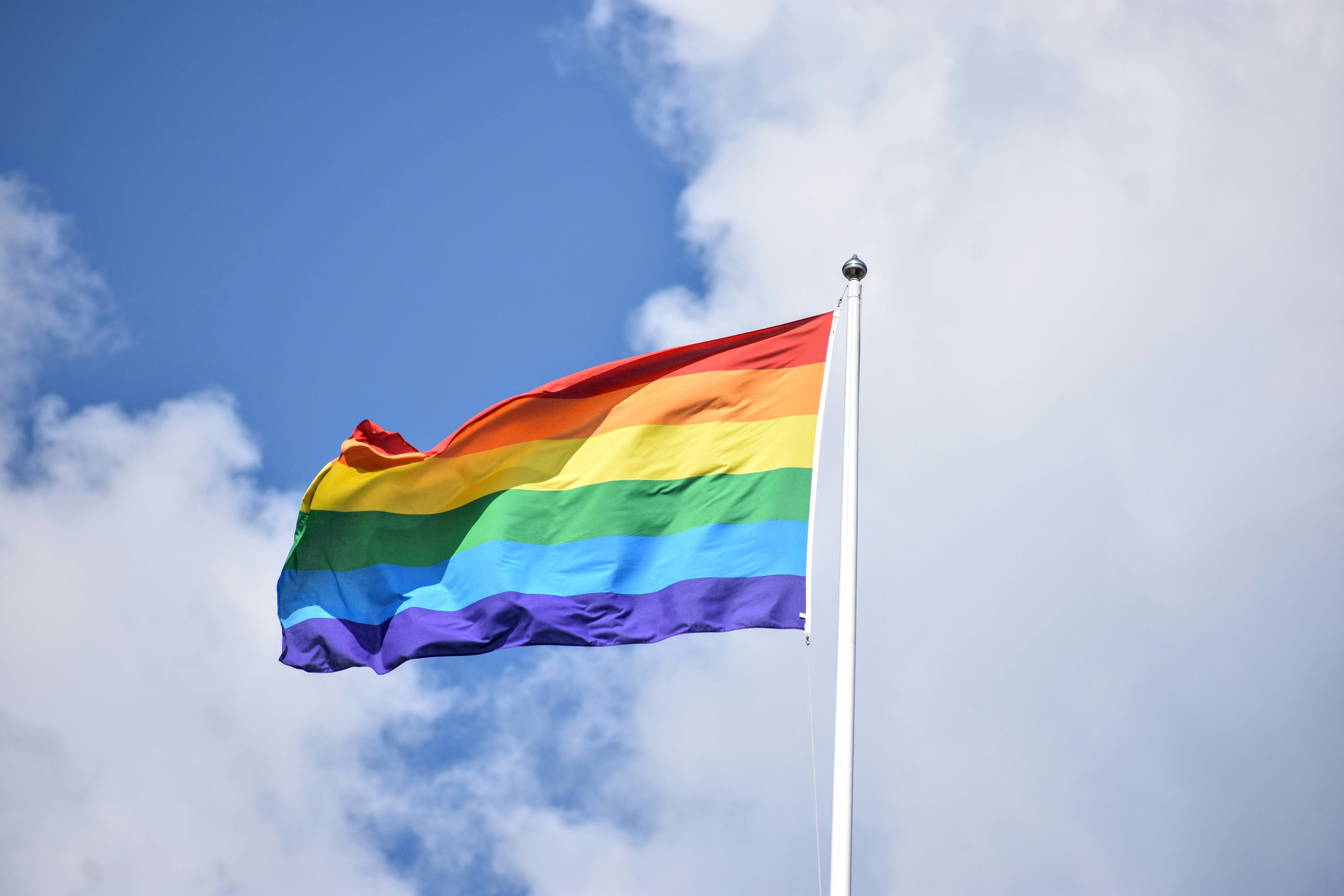 Zu sehen ist eine wehende Regenbogenfahne an einem Fahnenmast vor einem blauen Himmel mit weißen Wolken.