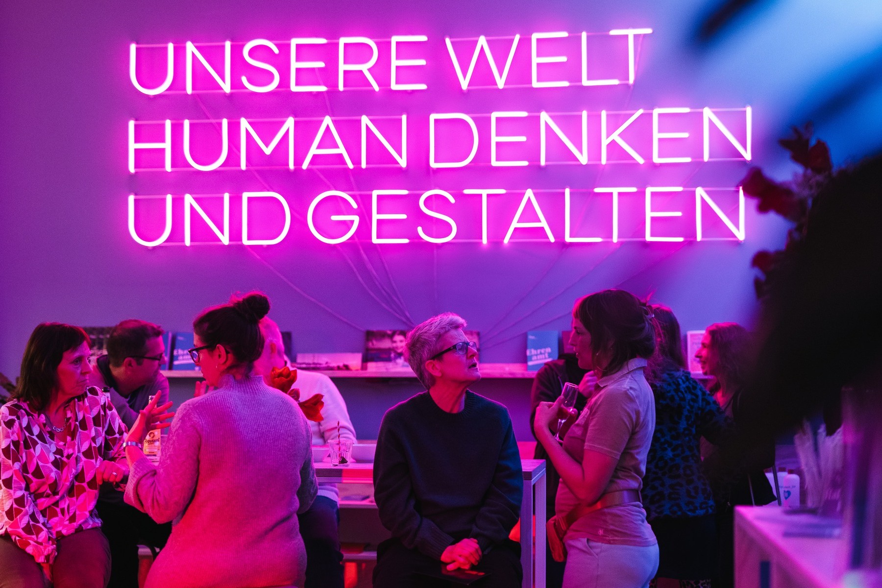 Im Haus des HUMANISMUS in der Potsdamer Straße in Berlin sind Leuchtbuchstaben an der Wand zu sehen. Zu lesen ist in Großbuchstaben: „Unsere Welt human denken und gestalten“. Davor sitzen Menschen an einem Tresen und unterhalten sich.